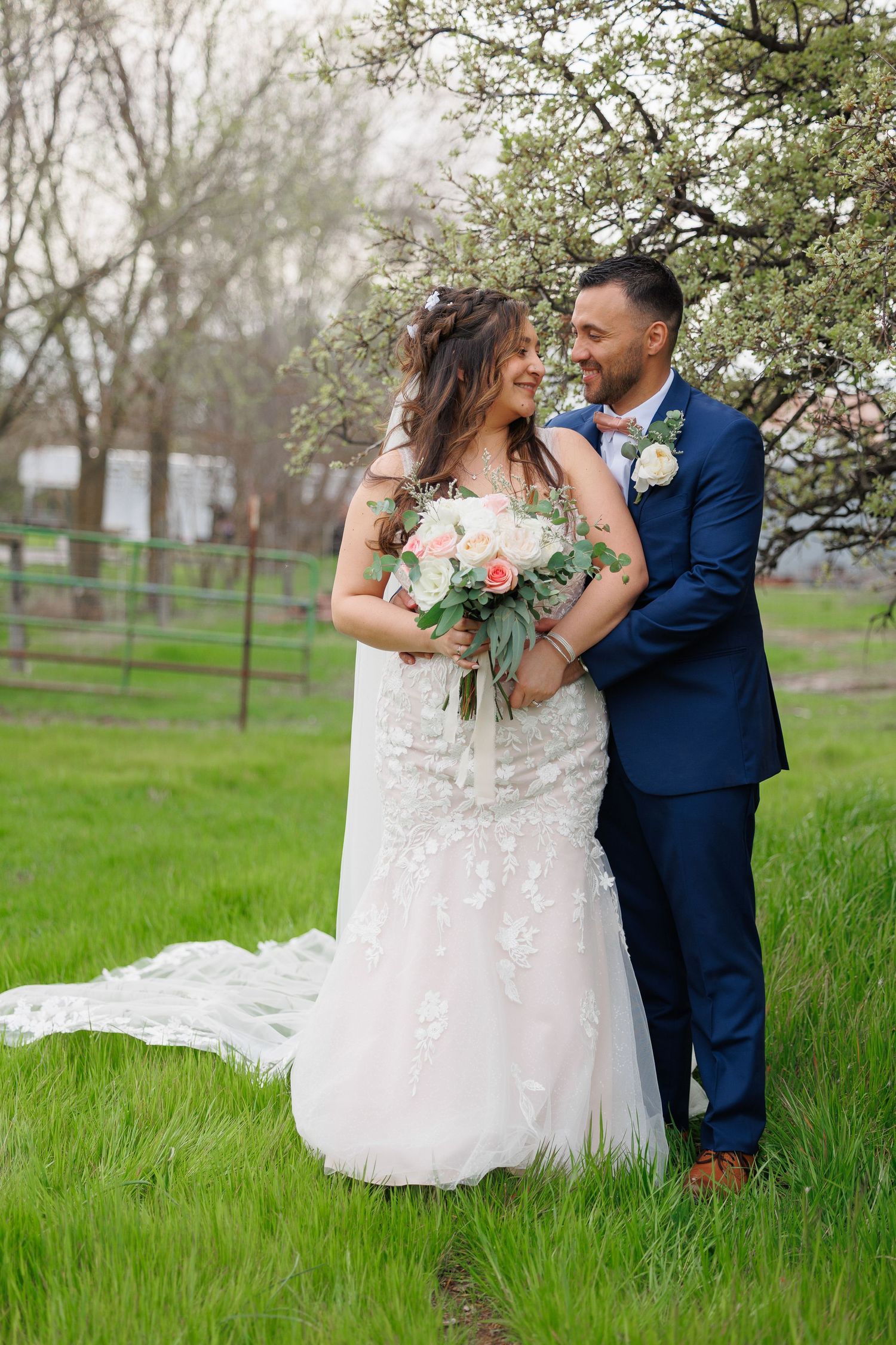 A couple in wedding attire embrace under a blooming spring tree in a lush green field.