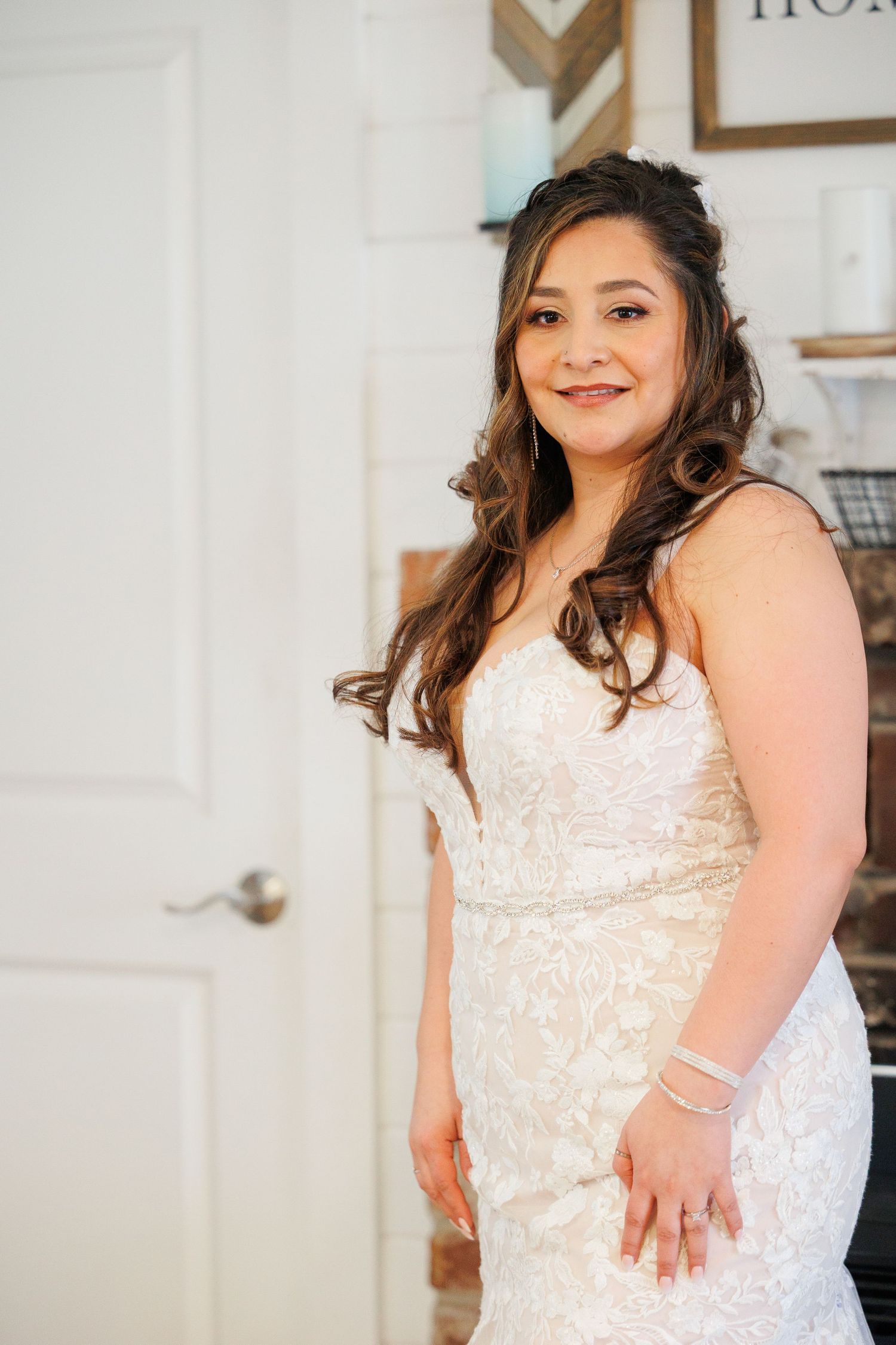 A person in a white lace dress poses against a light-colored wall with framed artwork in the background.
