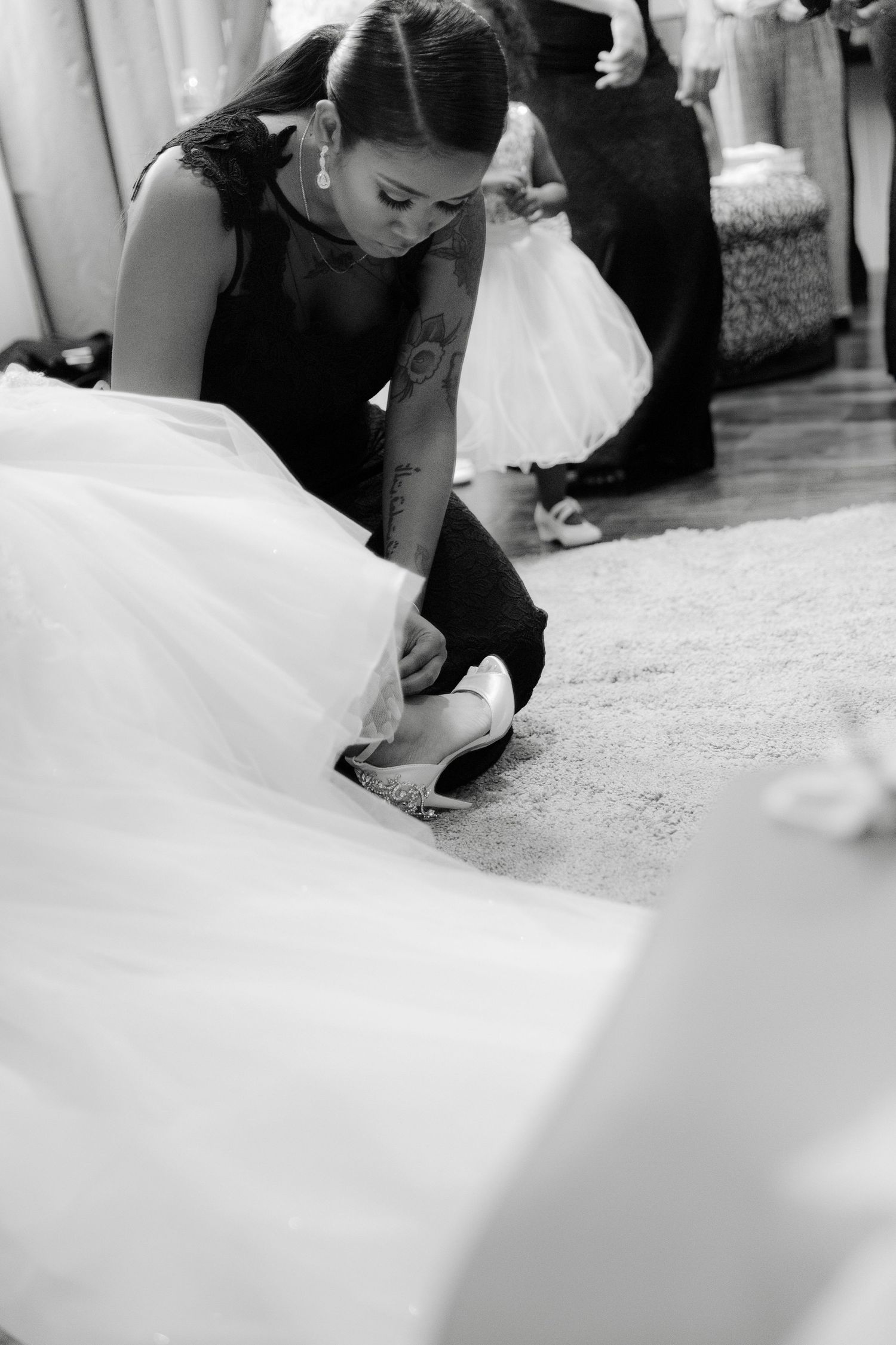 A person in black attire adjusts a white wedding dress and shoes on a light colored carpet.