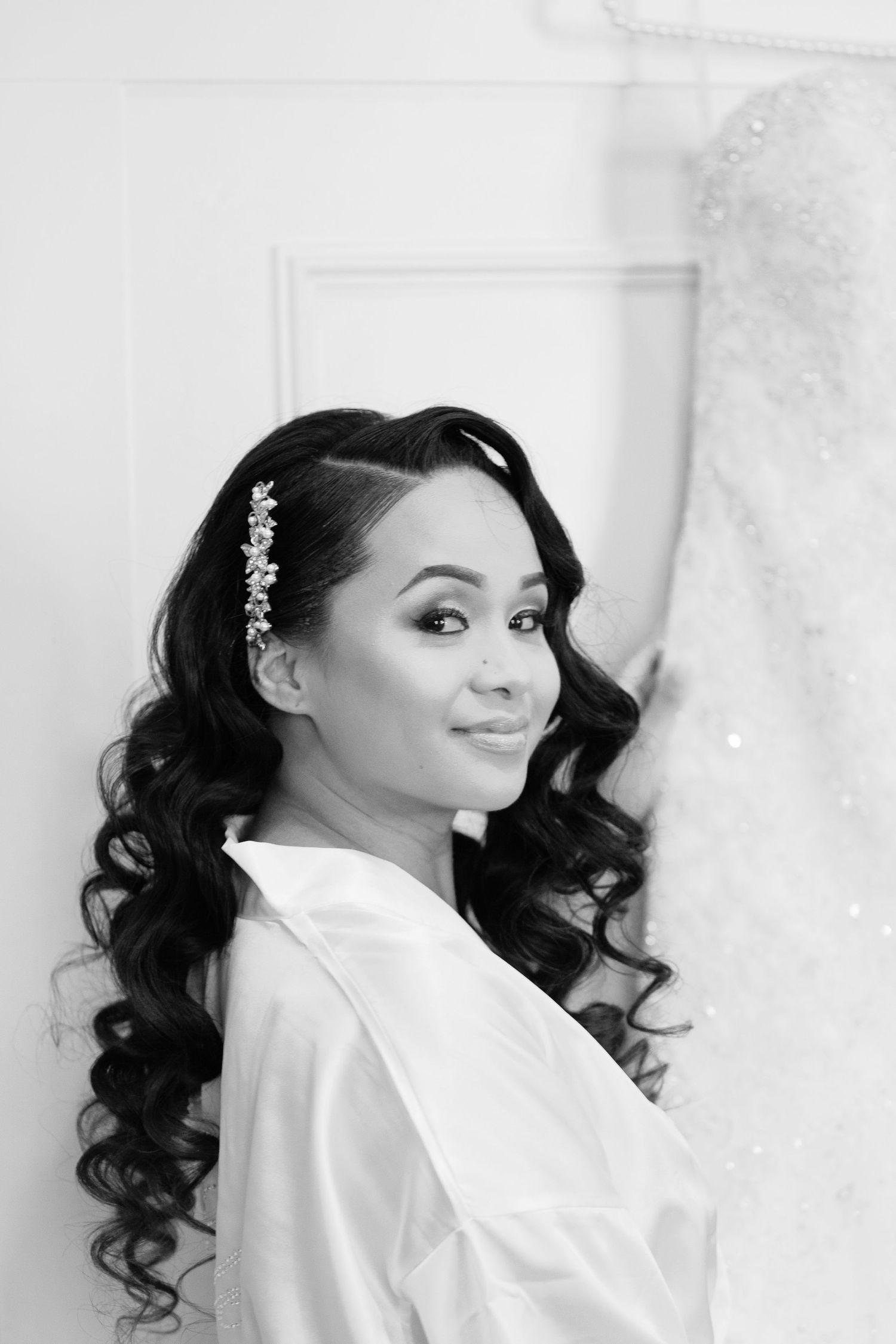 Black and white portrait of a bride with long curled hair and floral hair accessory wearing a white robe.