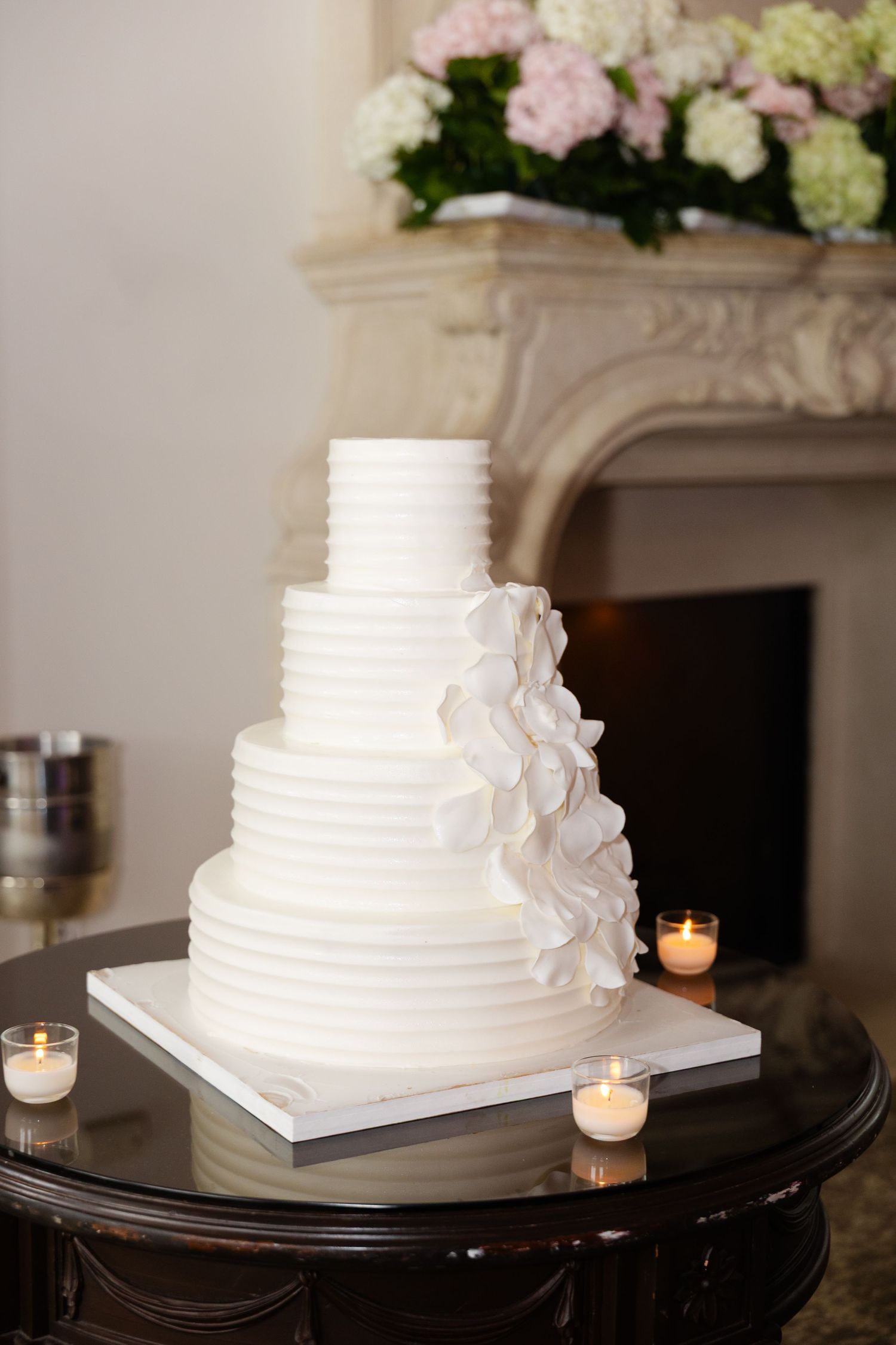 Elegant white tiered wedding cake with delicate ruffled details displayed on dark table with glowing votive candles.