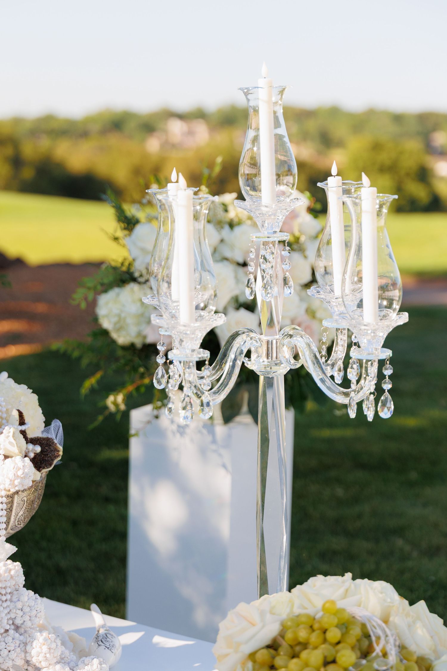 Elegant crystal chandeliers and white floral arrangements decorate an outdoor wedding reception setup at sunset.