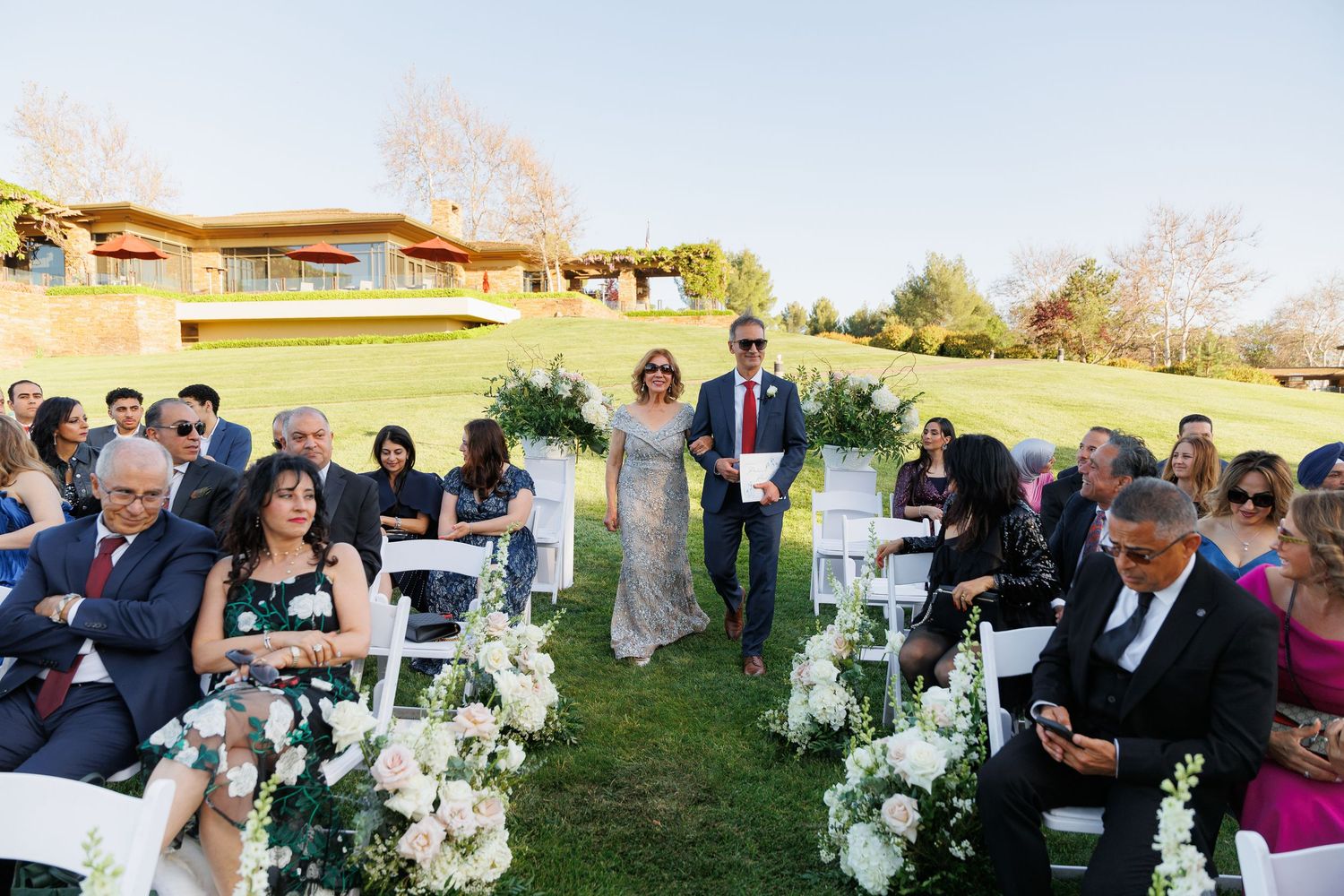 Wedding guests gather outdoors at a rustic venue with white floral arrangements and natural decor during sunset.