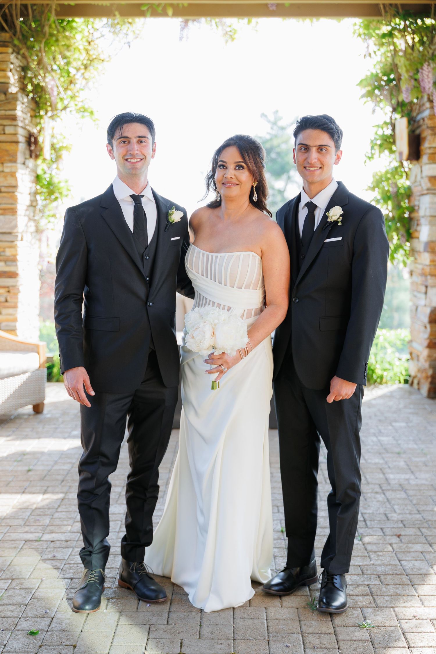 Wedding party in formal attire poses together on stone patio with black suits and white gown.