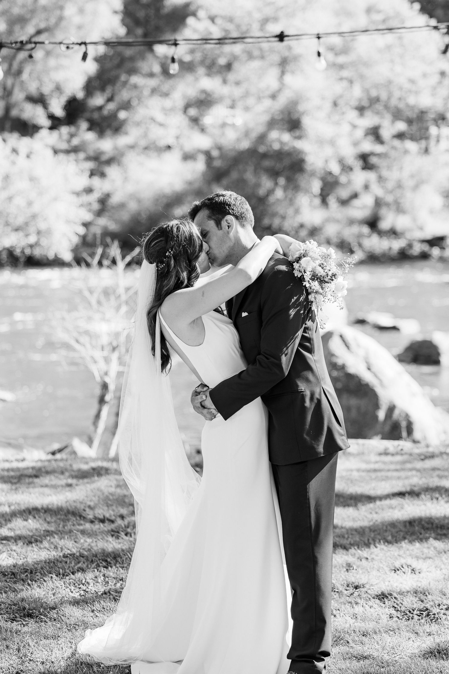 A romantic black and white wedding photo of a couple sharing a kiss outdoors with string lights and trees in the background.
