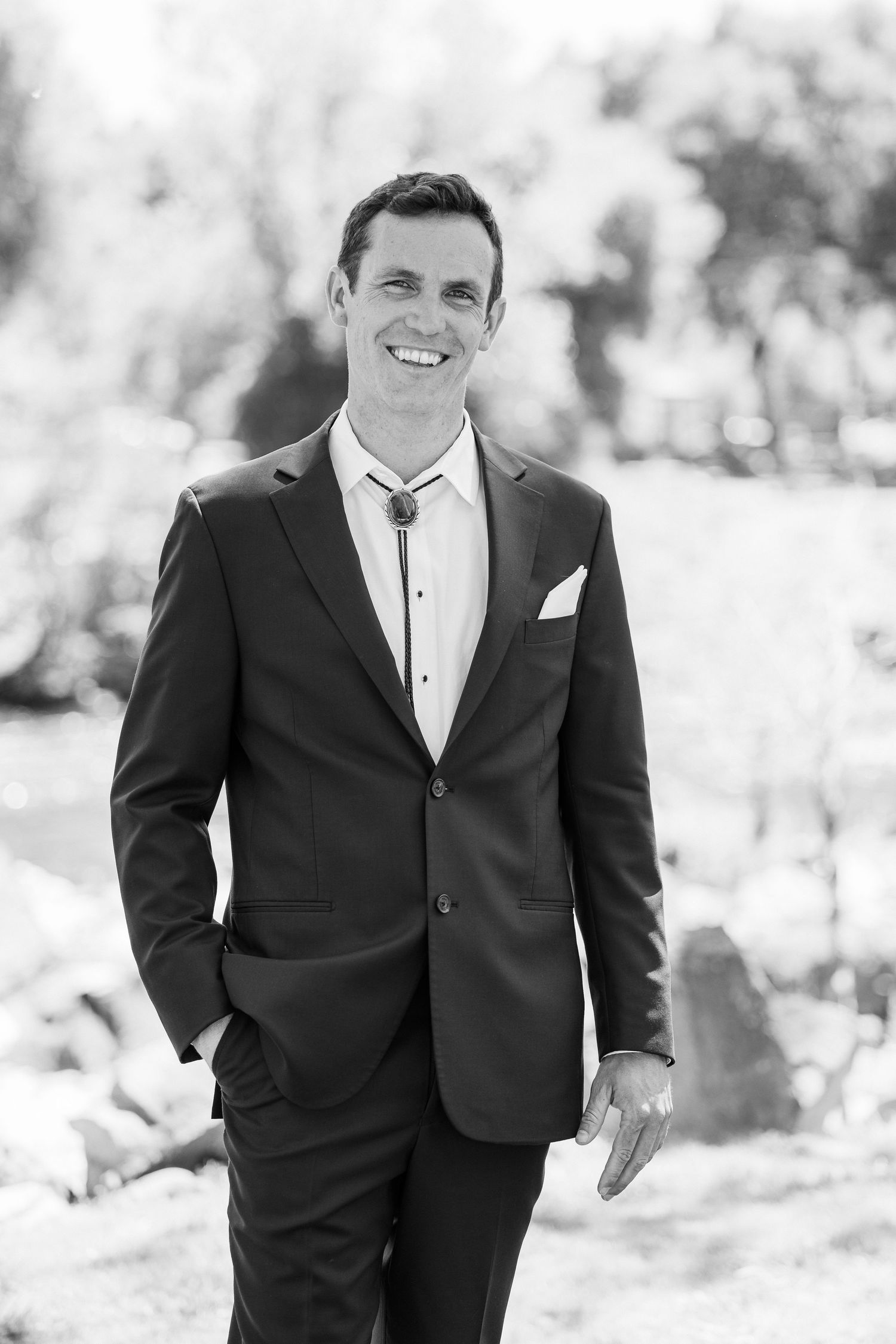 A black and white photograph of a groom in a formal tuxedo with white shirt and bow tie standing outdoors.