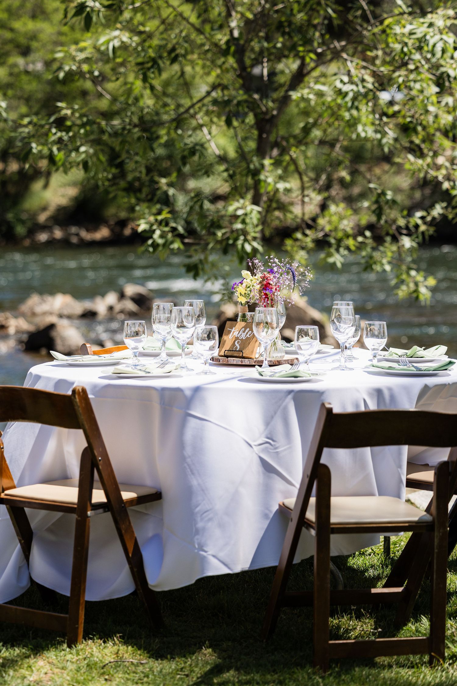 Elegant outdoor dining tables set with white linens and glassware along a riverbank under dappled sunlight and trees.