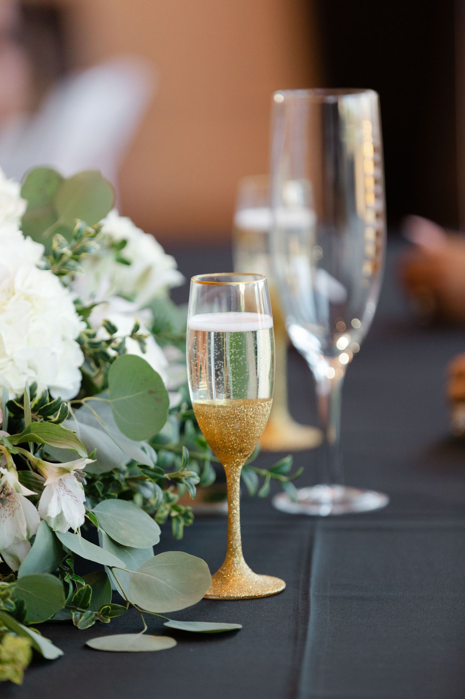Elegant champagne flute with gold glitter base sits beside white floral centerpiece on dark tablecloth.