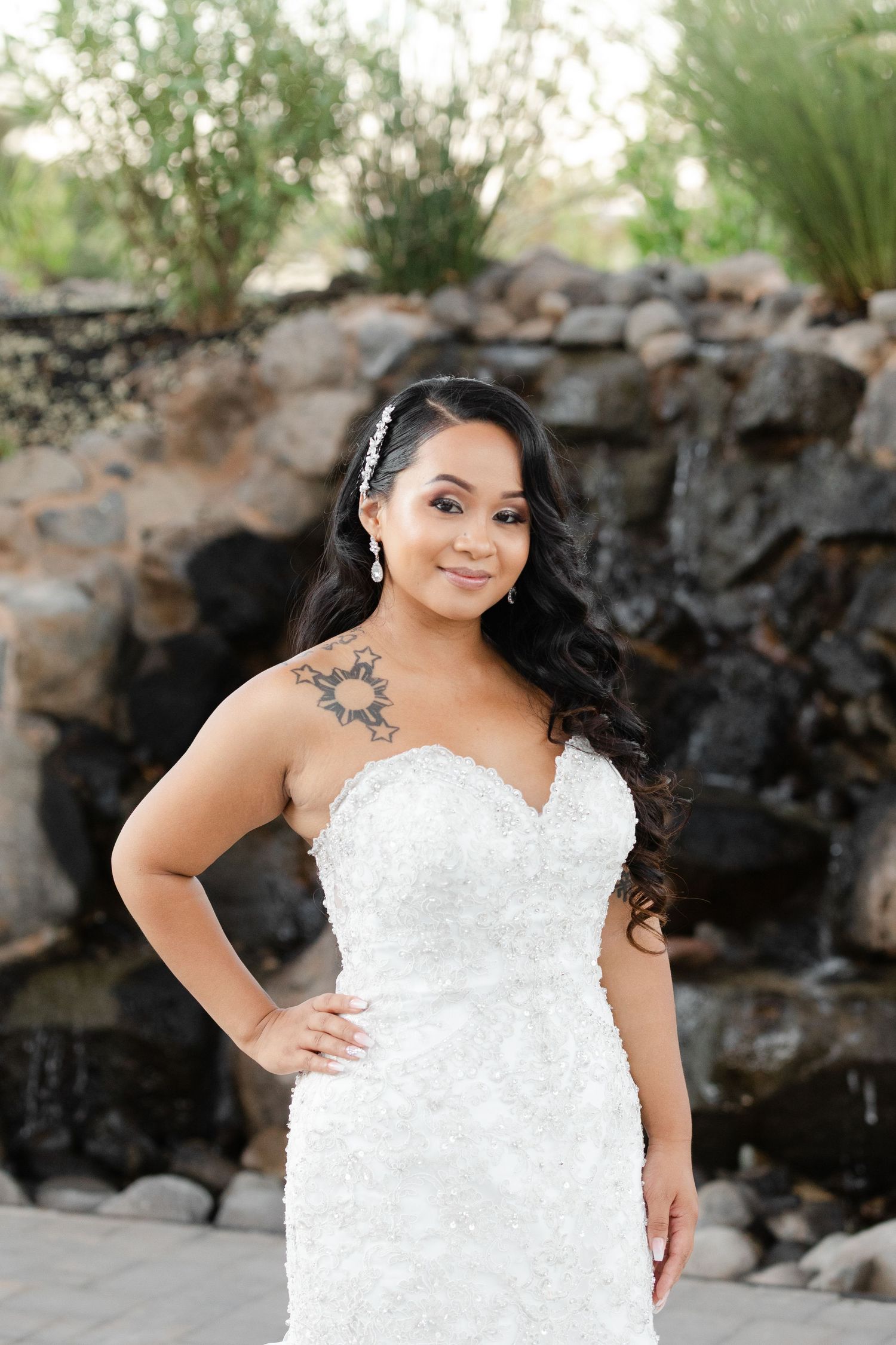 A bride in a white lace wedding dress poses against a natural rock wall backdrop in an outdoor setting.