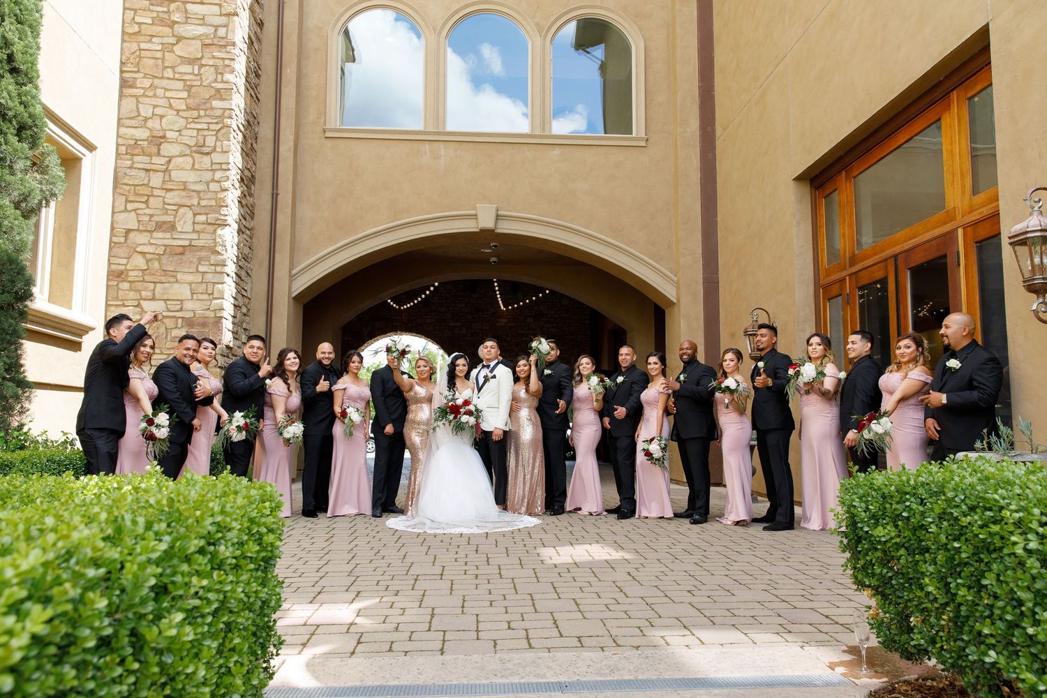 A wedding party in blush pink dresses poses together under an elegant stone archway entrance of a church.