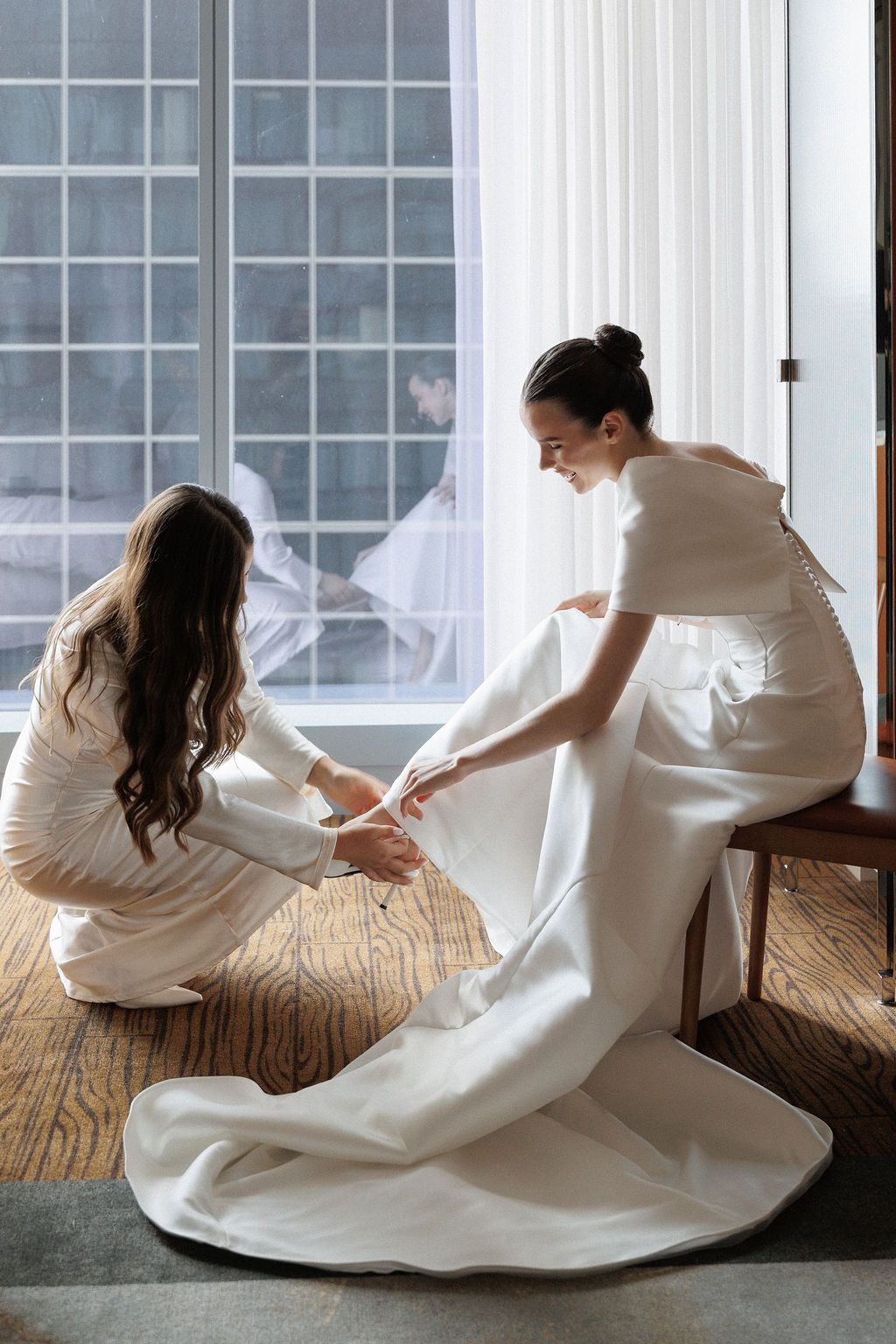 Two people in white dresses adjust wedding attire details near a bright window.