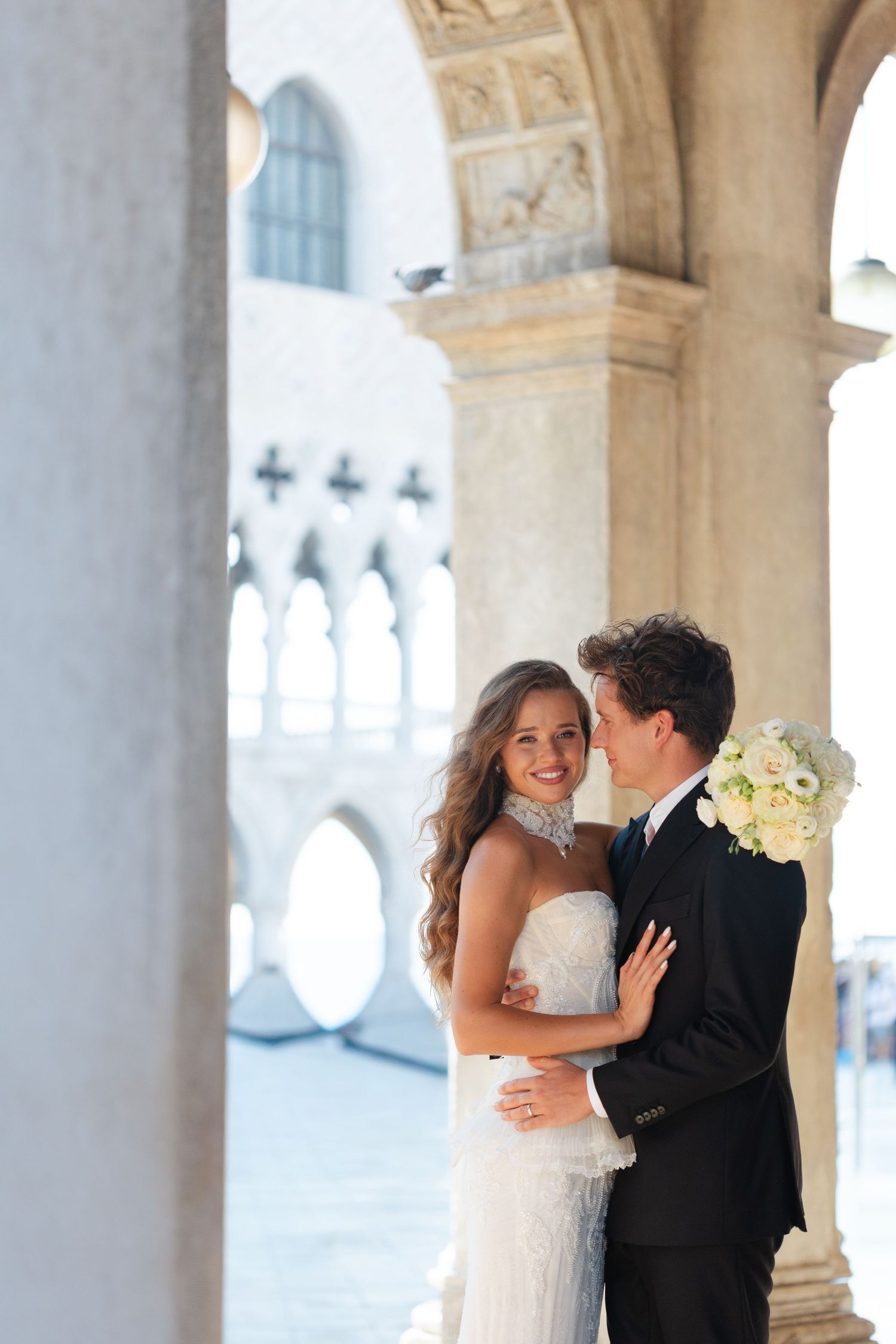 Newlywed couple embraces under ornate arches of Venice's Doge's Palace with white bouquet and elegant attire.