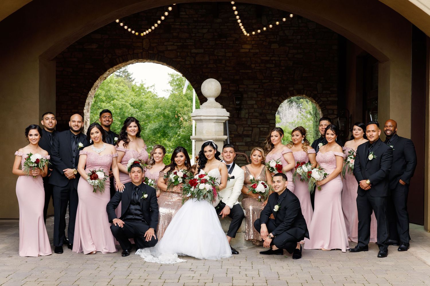 Wedding party in pink bridesmaid dresses and dark suits poses together on stone steps outdoors.