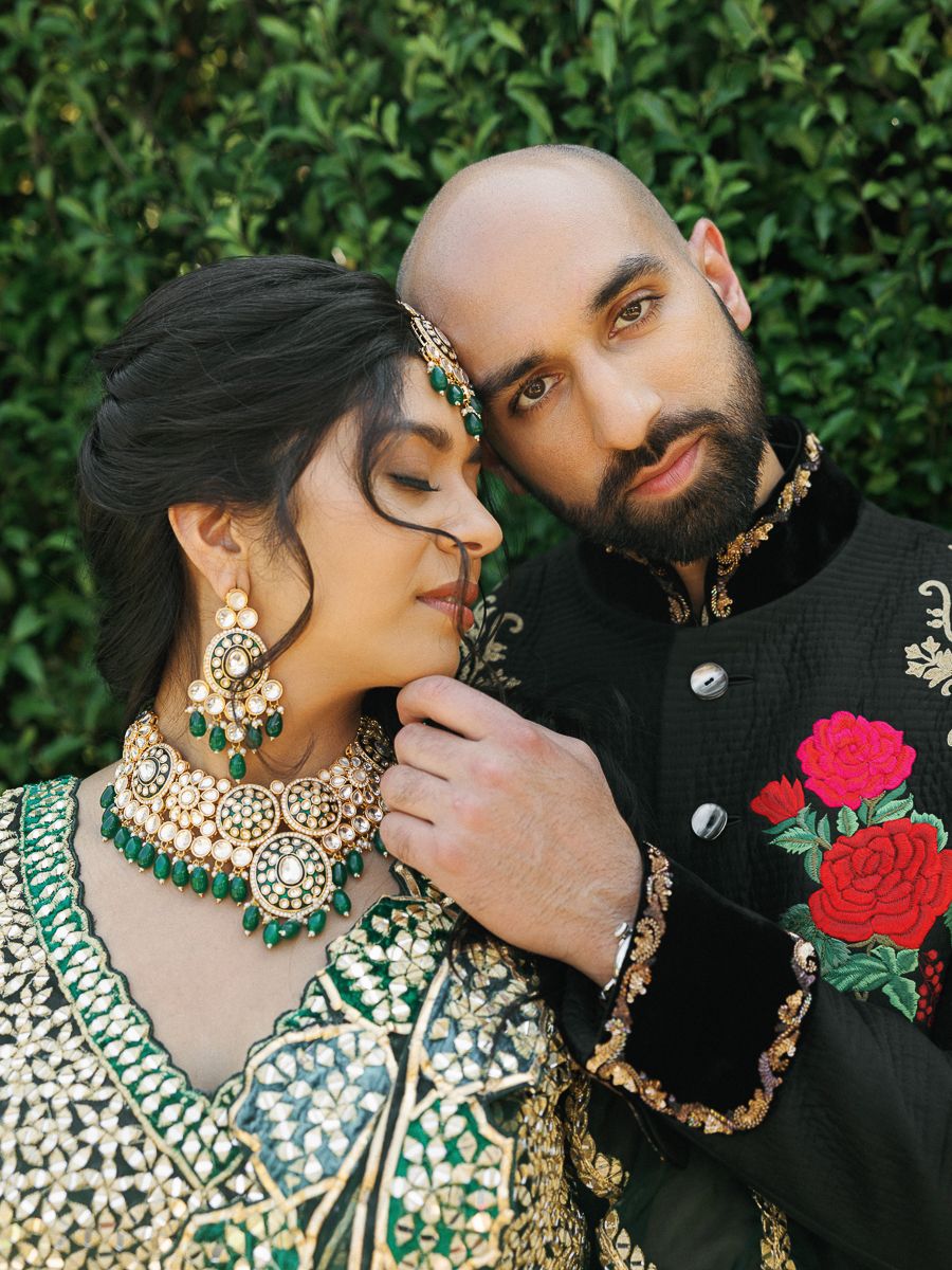 A romantic couple in traditional South Asian attire pose against a lush green hedge backdrop for their wedding photos.