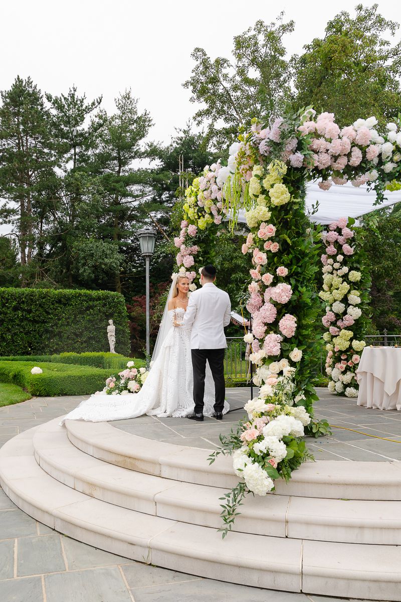 Wedding couple stands beneath an ornate floral arch with pink and cream roses on curved stone steps in a garden.