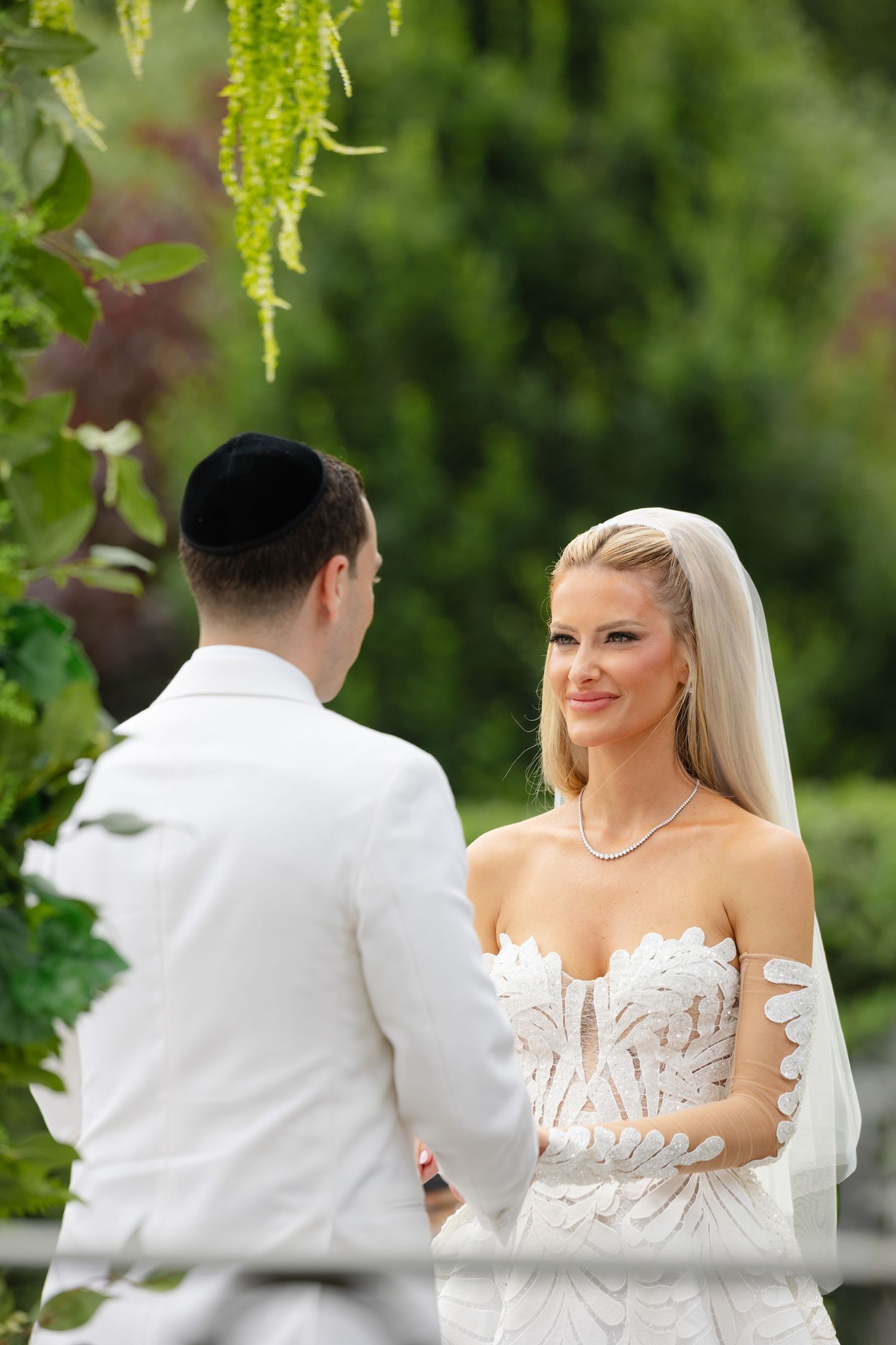 Bride in white floral gown smiles at groom wearing kippah during outdoor Jewish wedding ceremony.