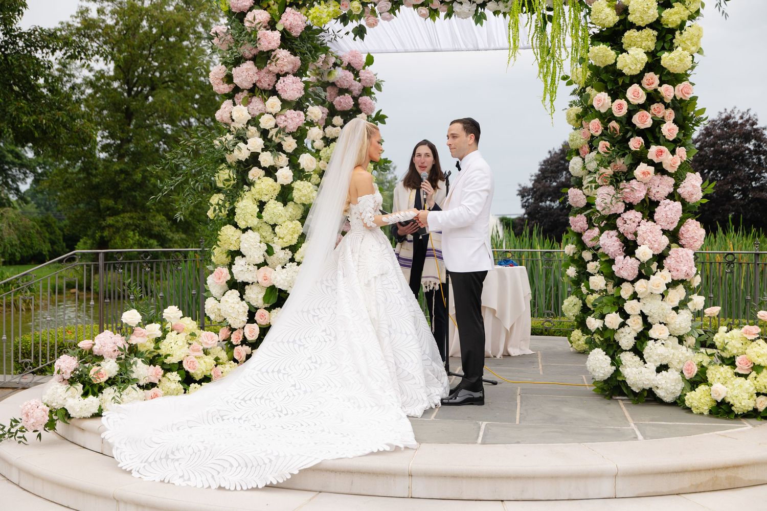 Couple exchanges vows under floral arch with pink and white roses at elegant outdoor wedding ceremony.