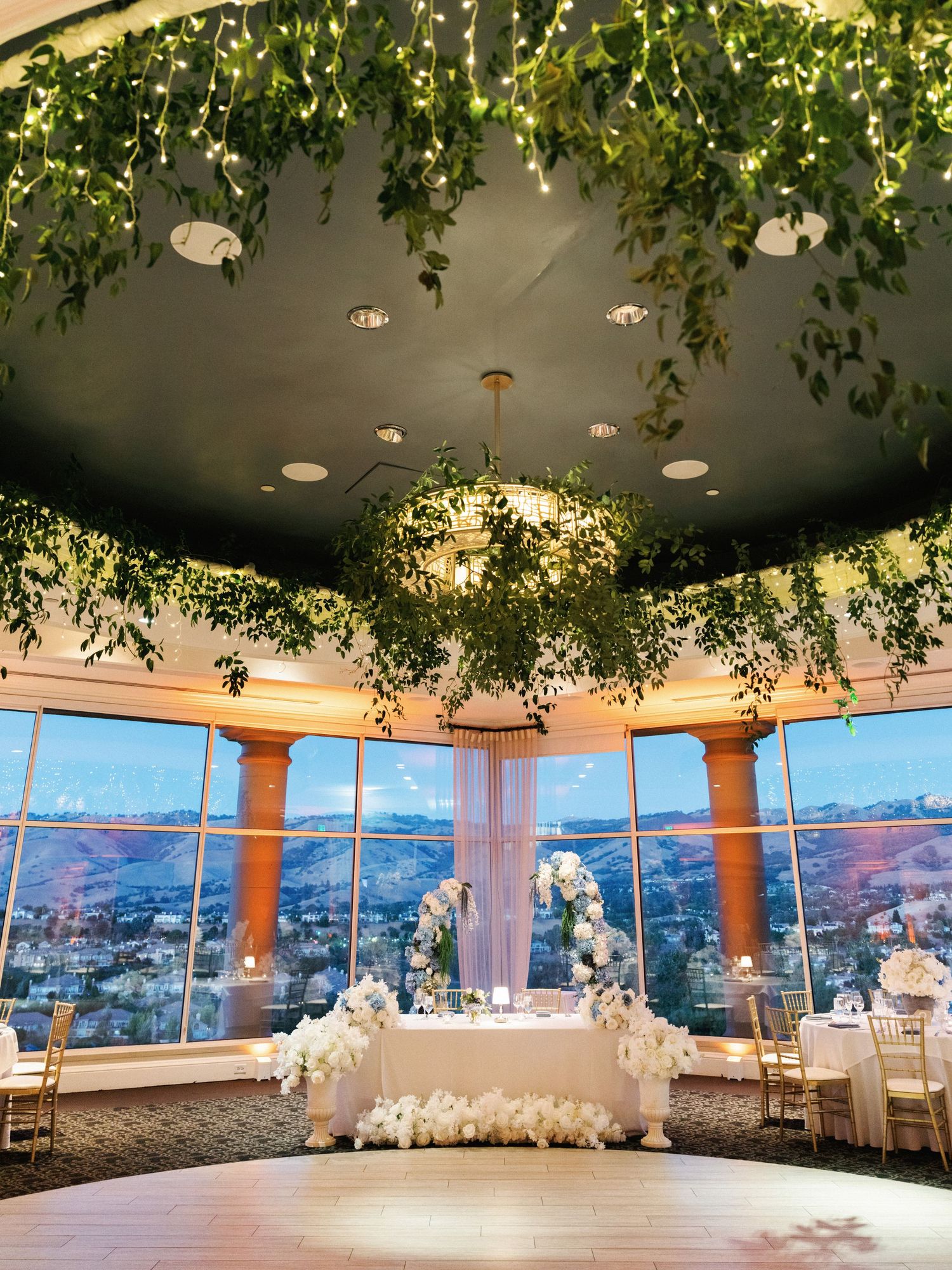 Elegant wedding reception venue with panoramic city views, white decor, and natural greenery hanging from the ceiling.