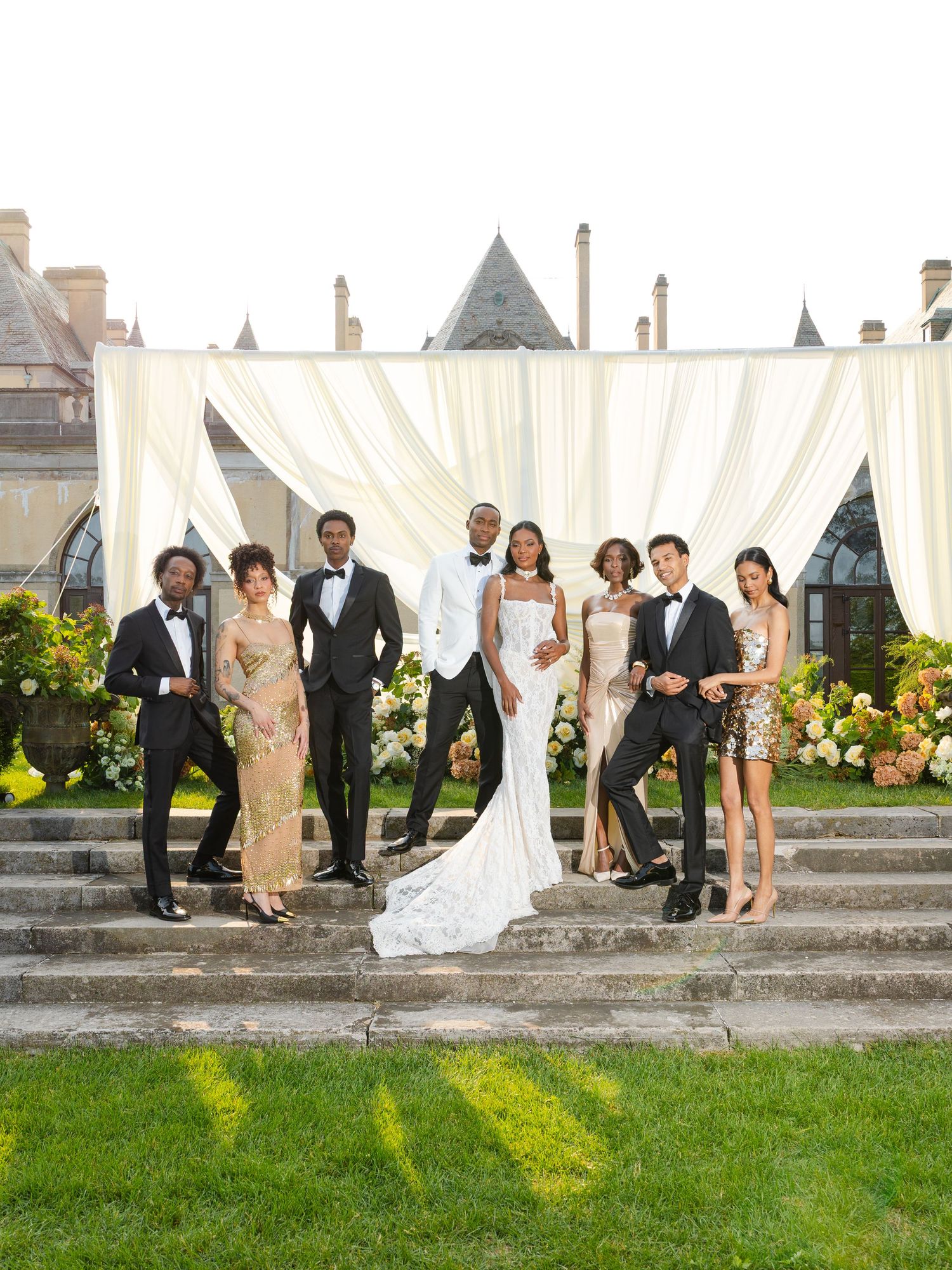 Wedding party poses against stone wall with white tent and colorful floral arrangements in outdoor venue.