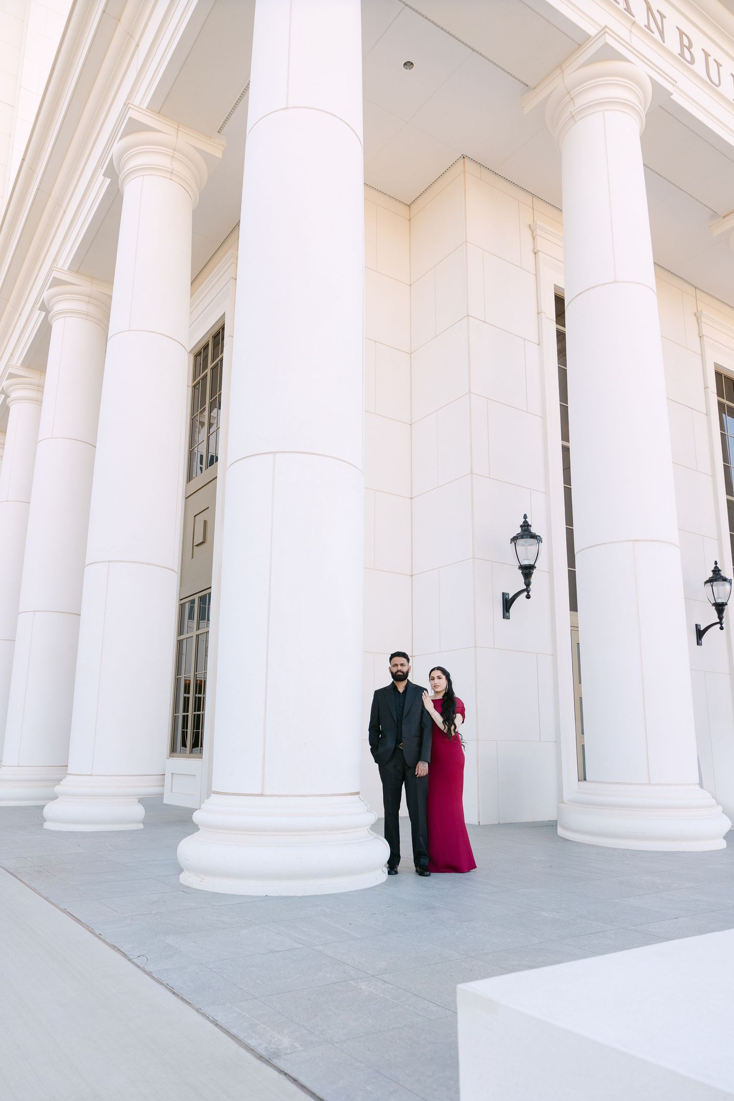 Elegant couple poses between grand white columns of a classical building with the woman wearing a flowing red gown.