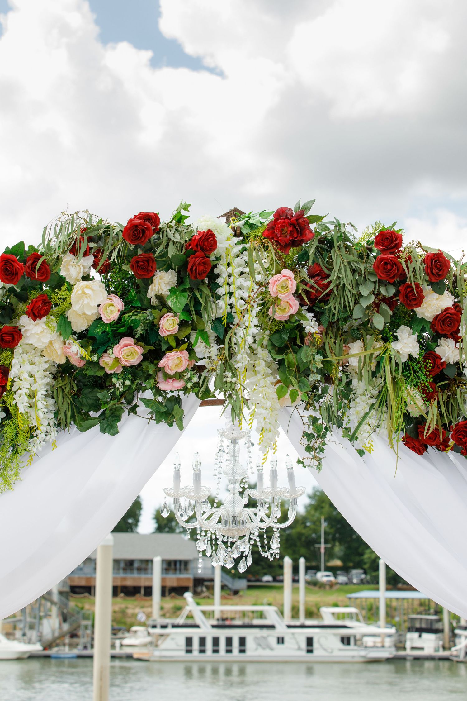 Elegant white wedding arch decorated with cascading red and pink roses, greenery, and crystal chandeliers over a waterfront view.