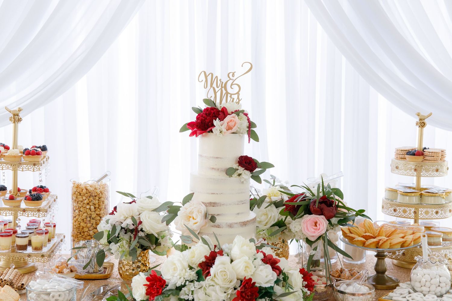Elegant white tiered dessert stand with pastries and floral decorations in burgundy and pink against a draped backdrop.