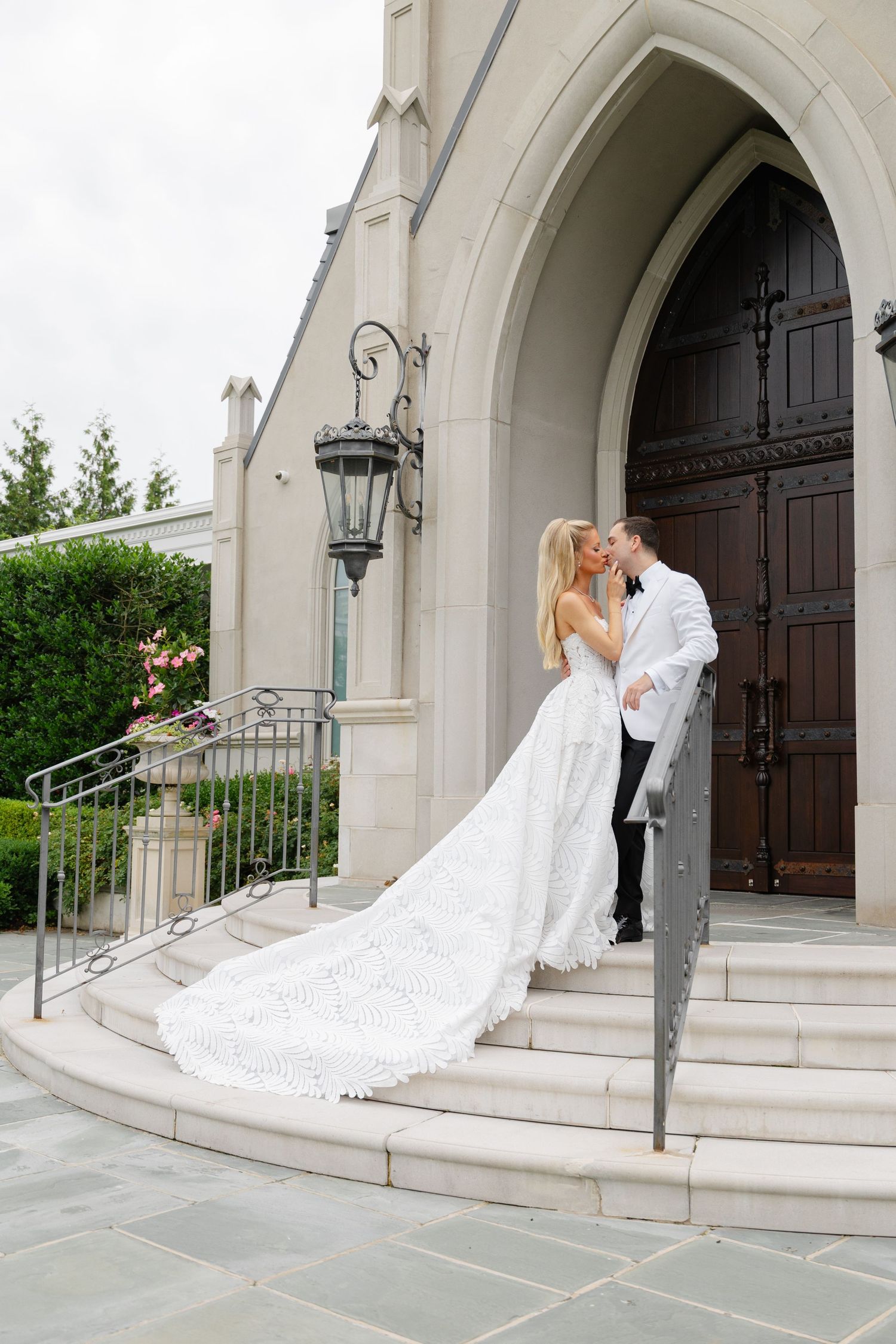A romantic wedding couple shares a kiss on the steps of a grand church with stone archways and wooden doors.