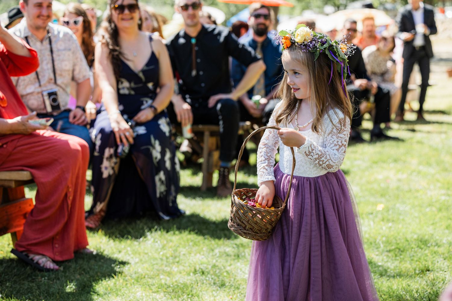 A flower girl in a purple dress walks down a grassy aisle at an outdoor wedding ceremony.
