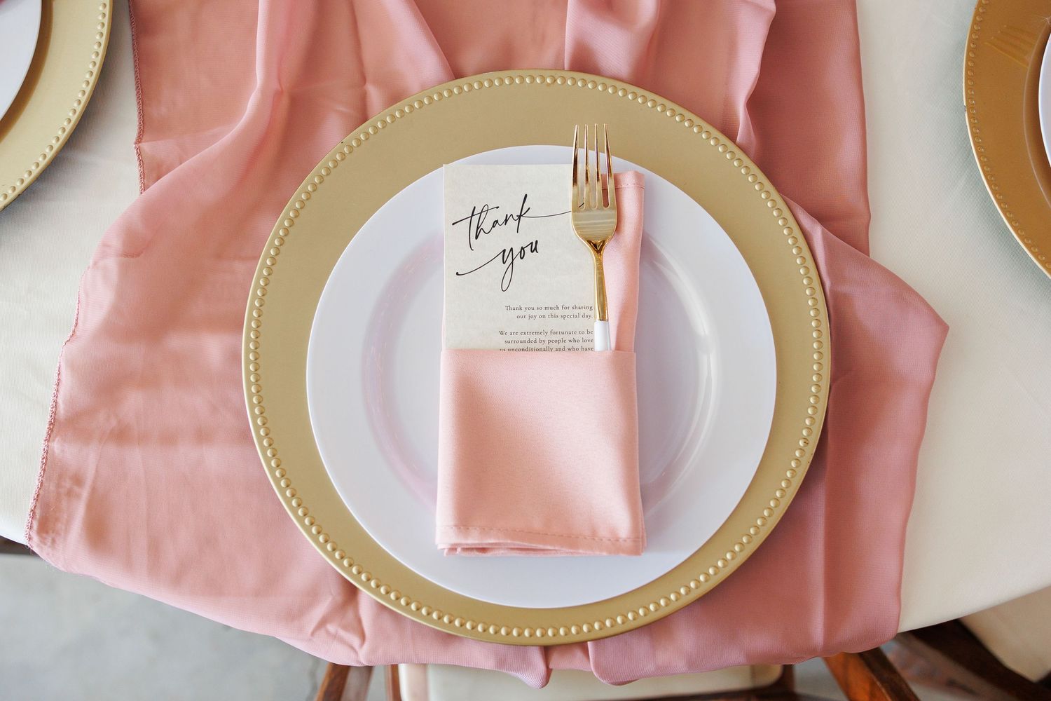 Elegant place setting with gold-rimmed charger plate and pink linen napkin on light pink tablecloth.