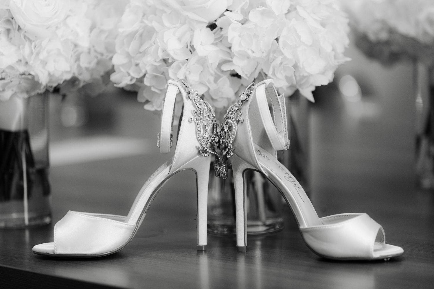 Elegant white bridal heels displayed next to white flowers in a black and white photograph.