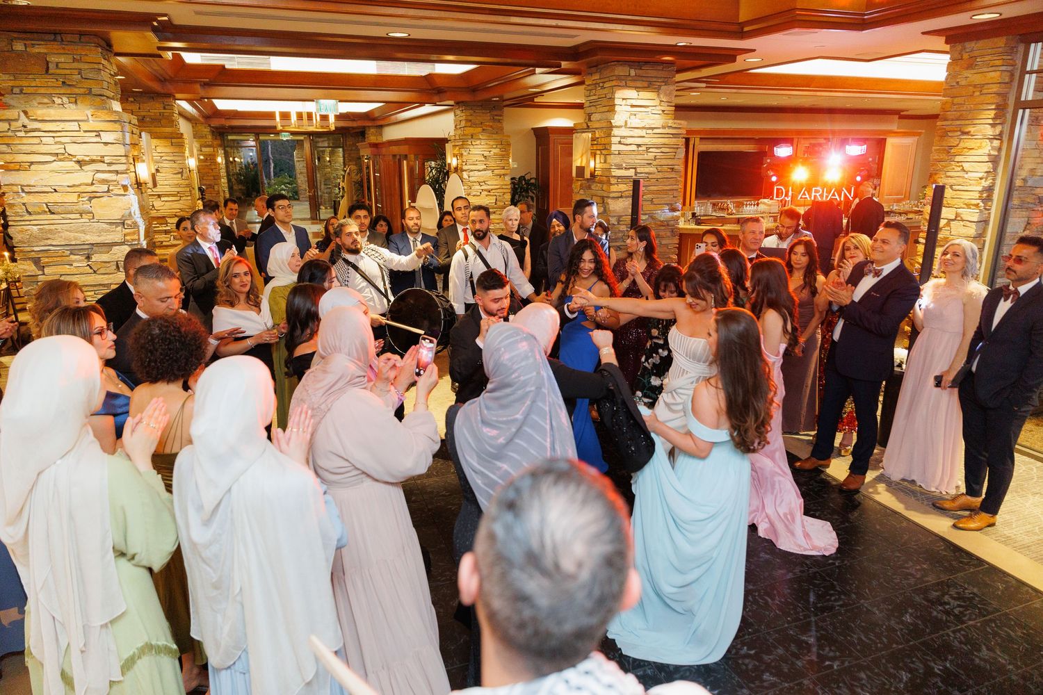 Wedding guests dance together during a reception celebration in a rustic stone-walled venue with warm lighting.