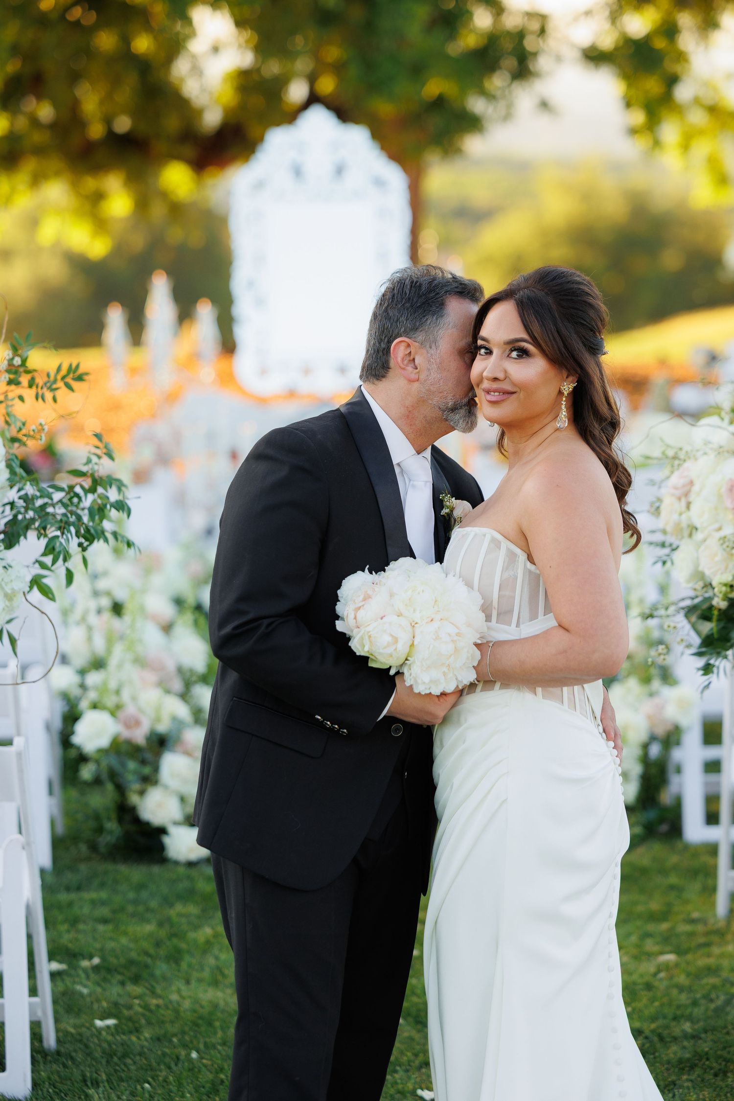 A romantic wedding couple shares intimate moments in a garden setting with white floral decorations at sunset.