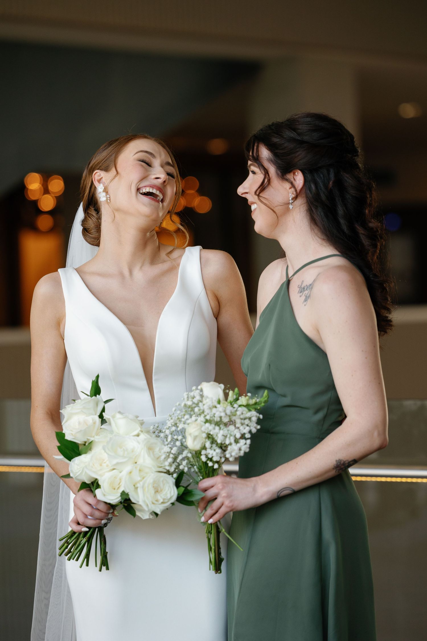 Wedding party members in white and sage green dresses share candid laughing moments while holding white bouquets.