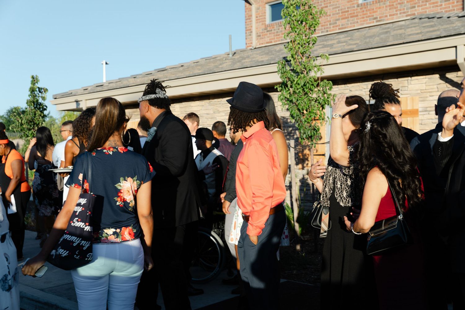 A long line of people wait outside a brick building during the daytime.