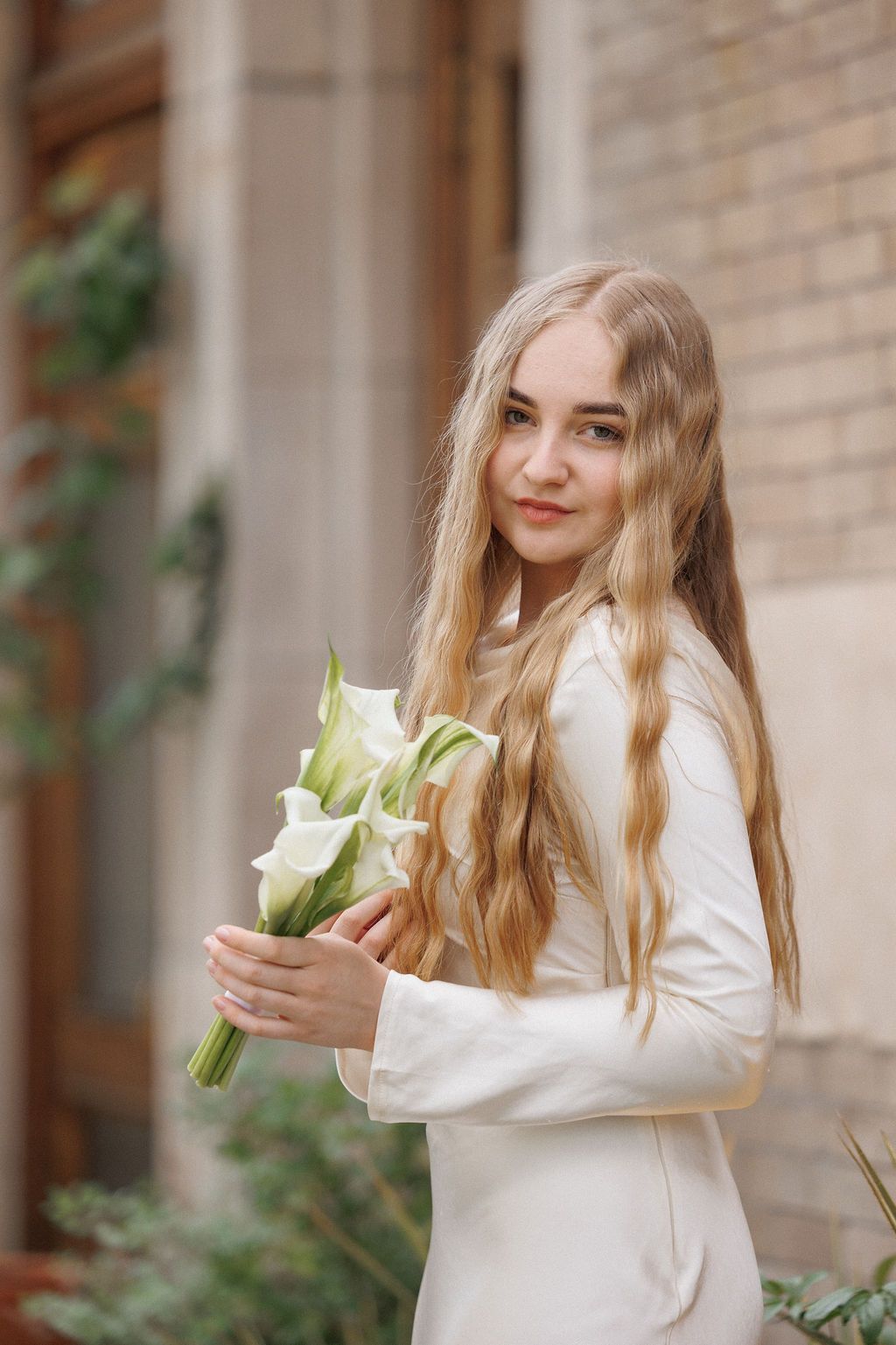 Person in white dress holds calla lily flowers against brick wall with climbing vines.