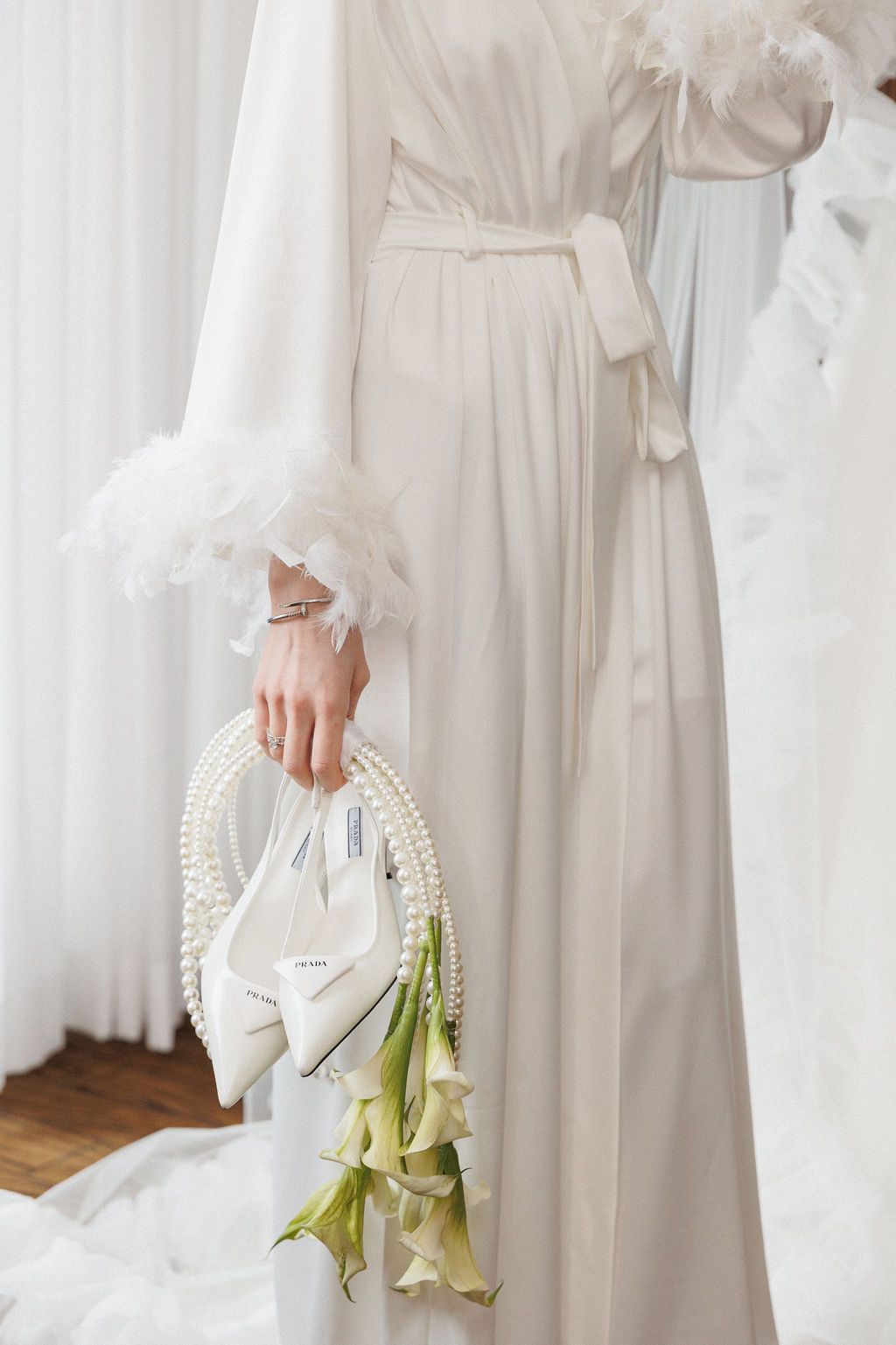 Woman in white vintage-style dress holds delicate pearl-beaded basket with green floral stems against light backdrop.