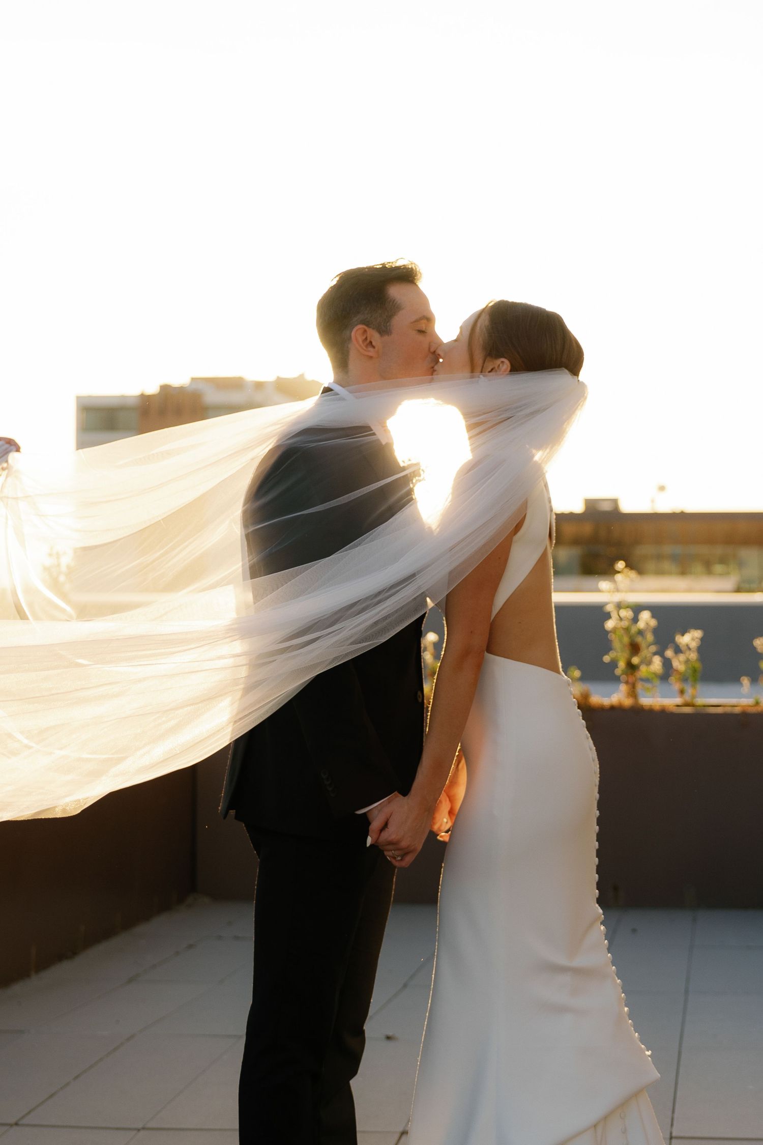 A romantic wedding couple stands together as a flowing veil catches the golden sunlight during sunset.