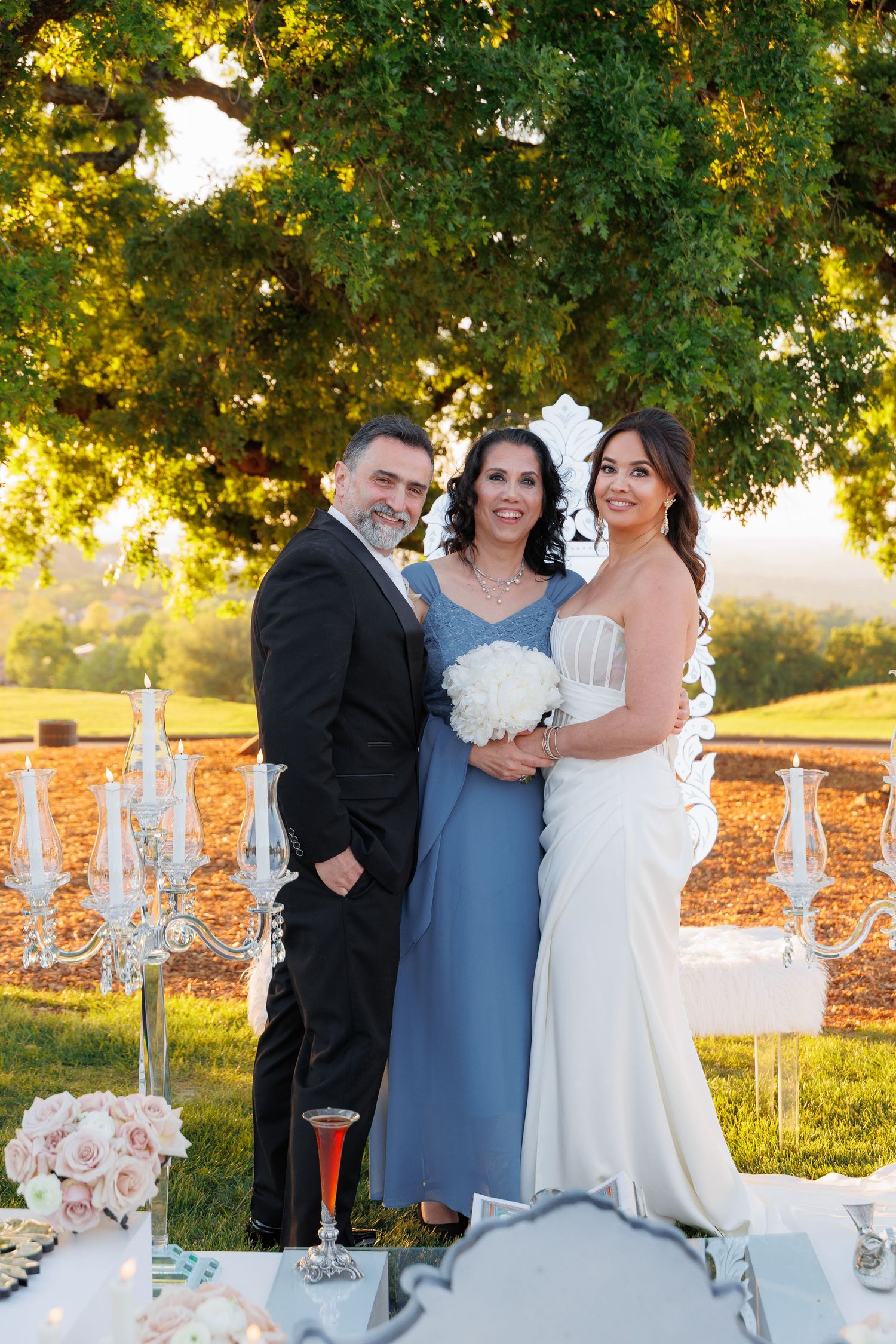 Wedding celebration outdoors with elegant table settings and crystal candelabras under a golden sunlit tree.