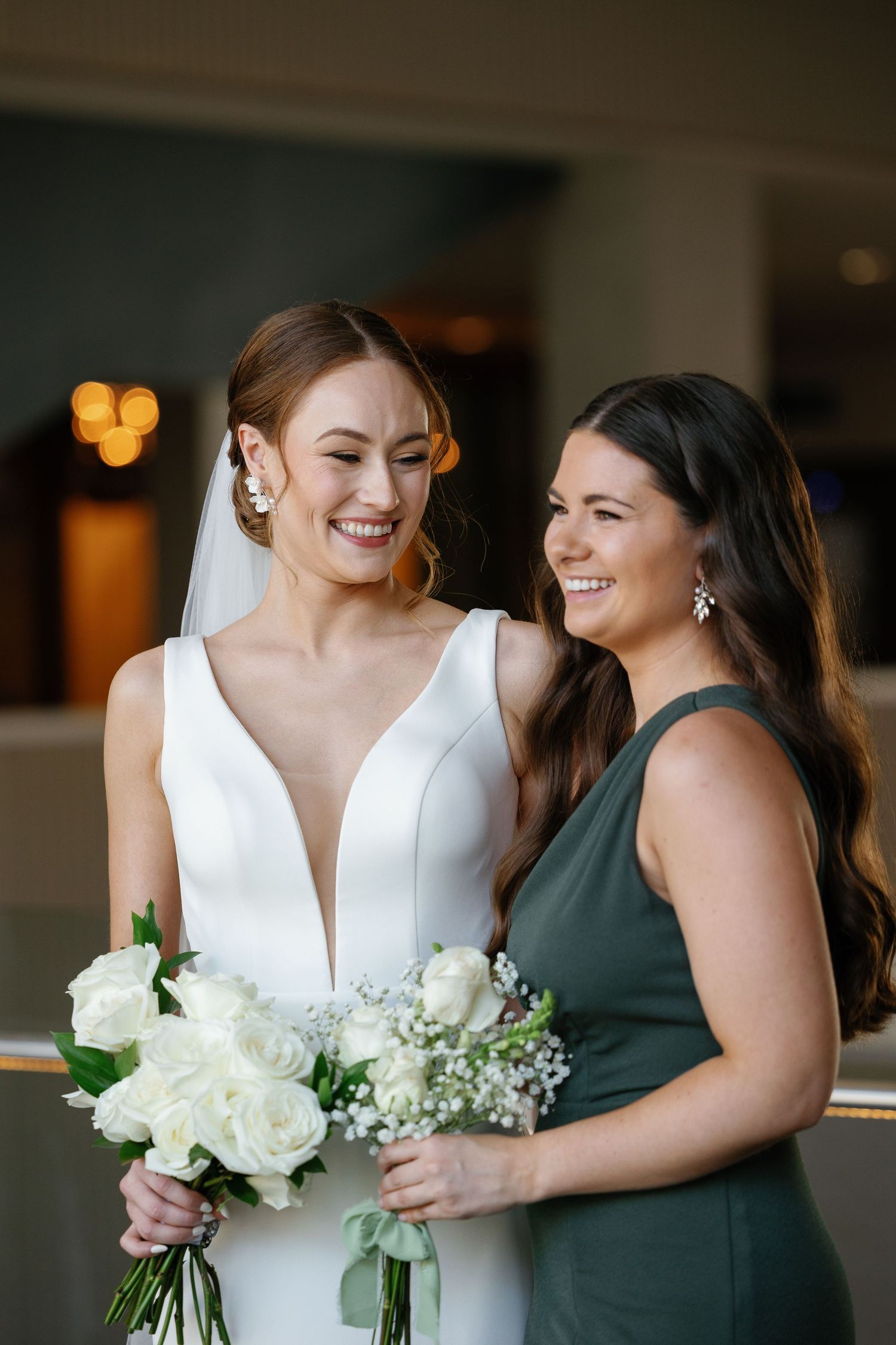 A bride in a white v-neck gown and bridesmaid in dark green dress share a joyful moment while holding white bouquets.