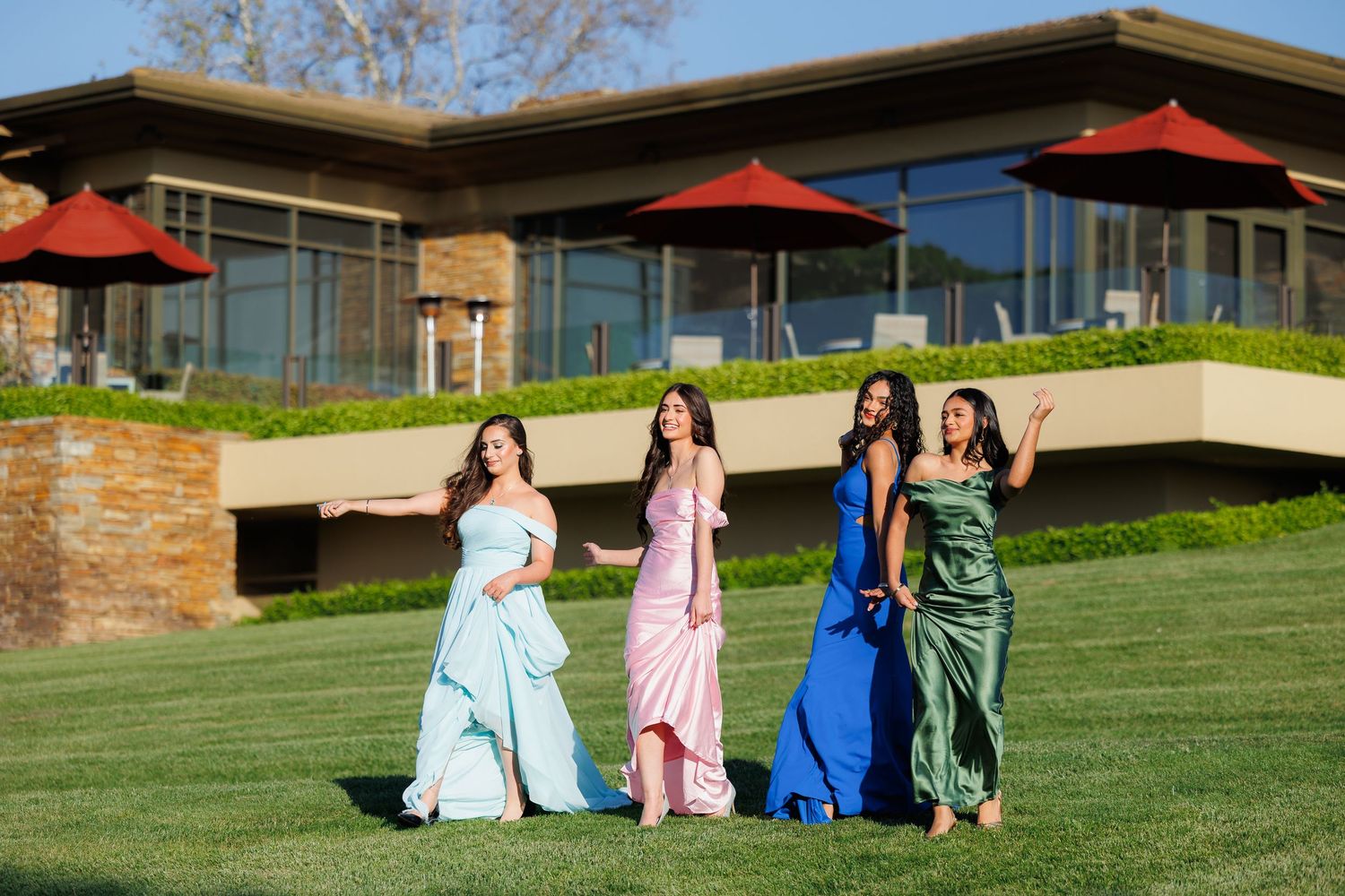 A group in colorful formal dresses poses together on a grassy lawn with red umbrellas and modern buildings in the background.