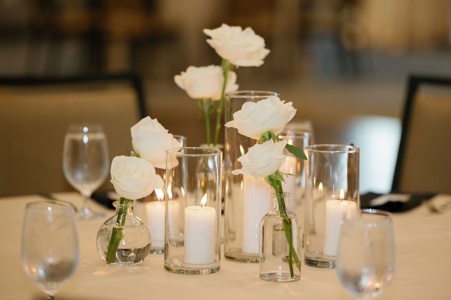Elegant wedding reception table settings with white roses in bud vases and glowing votive candles.