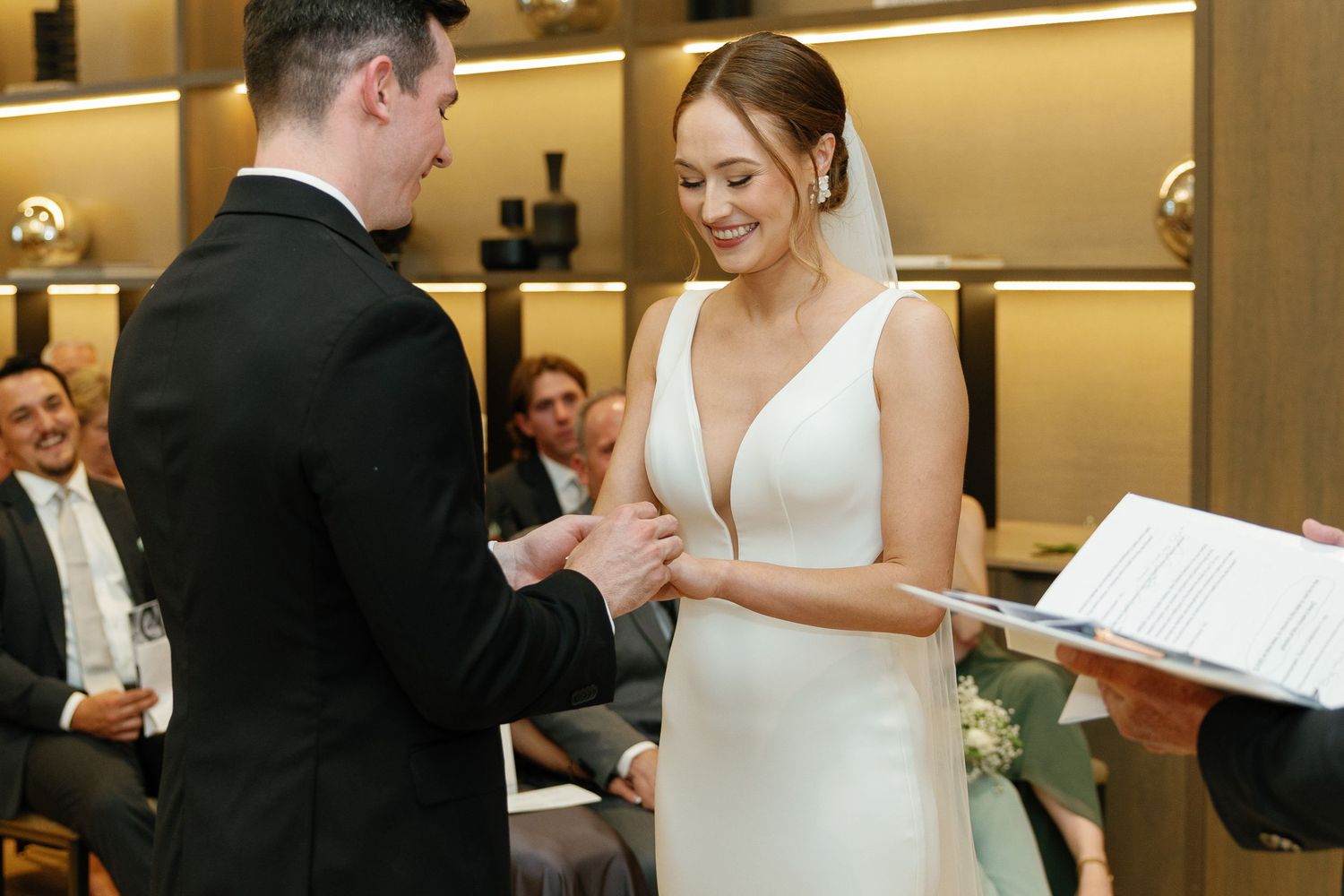 A couple exchanges rings during an intimate indoor wedding ceremony while guests look on.