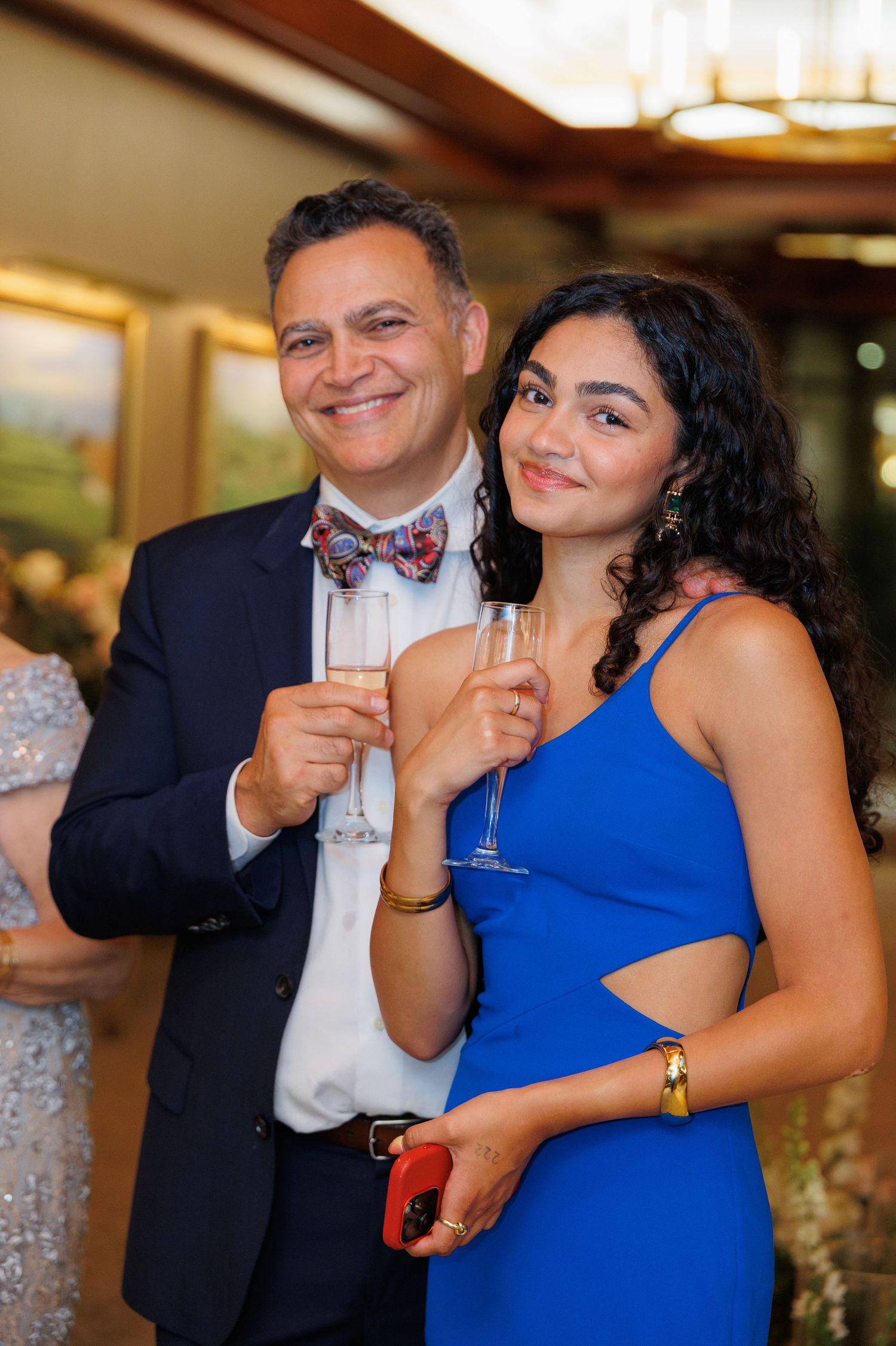 A couple shares a playful moment with drinks at an elegant event, with the woman wearing a vibrant blue dress.
