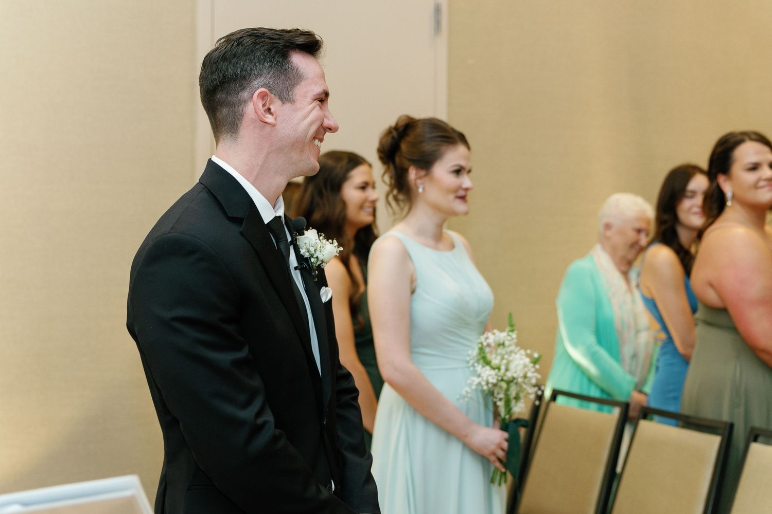 Wedding party members stand in a line wearing formal attire including tuxedo and pastel bridesmaid dresses.