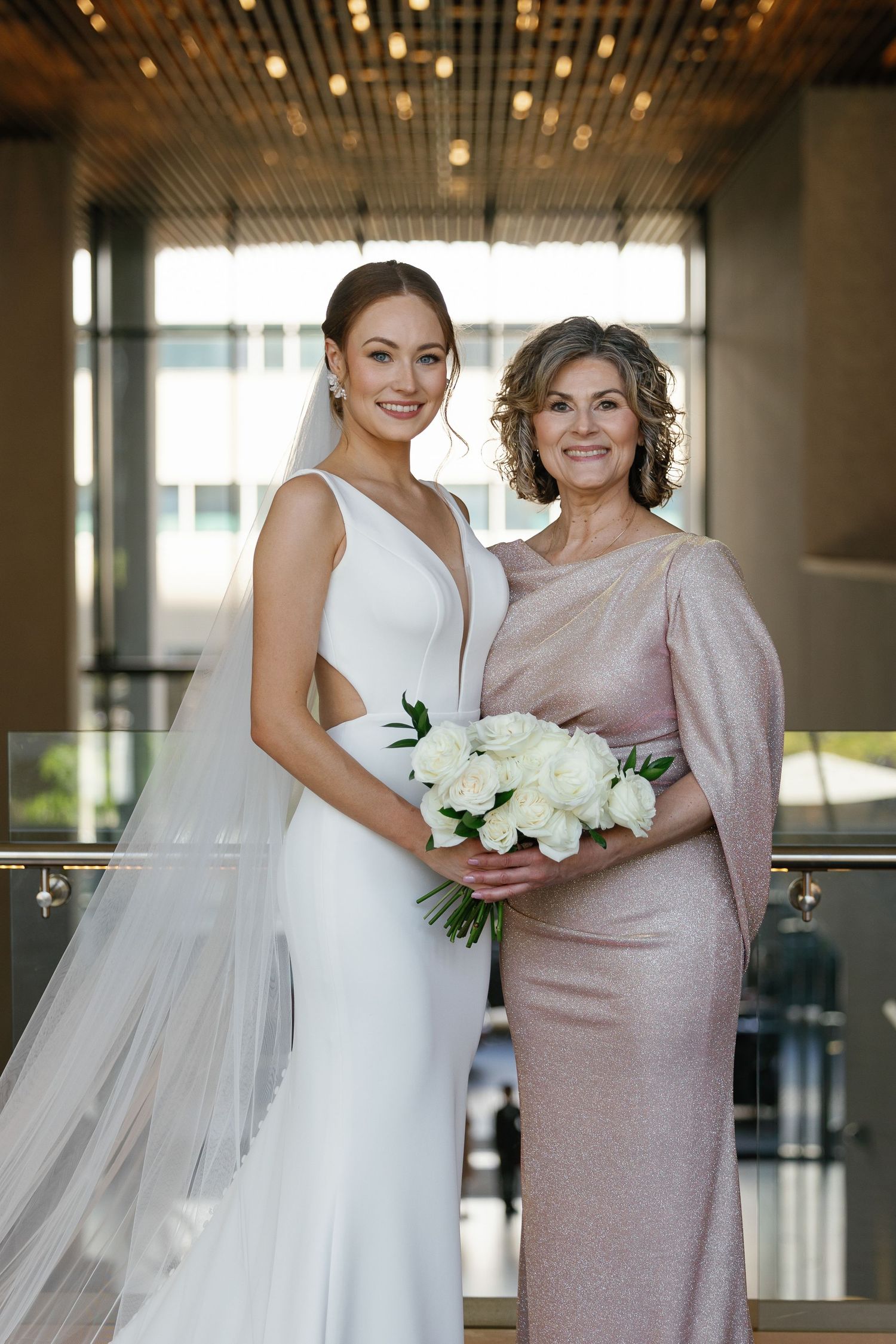 Two people in formal attire share a tender moment, with one wearing a white wedding dress and holding white flowers.
