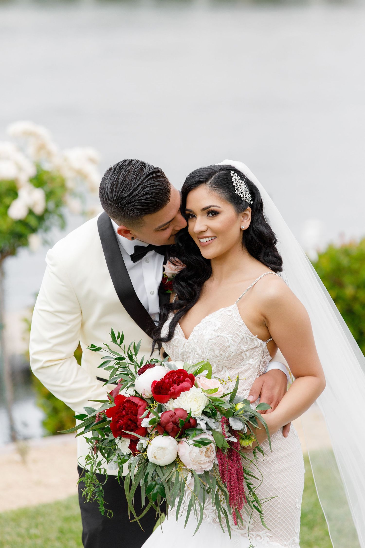 A romantic wedding couple embraces outdoors with a stunning red and white floral bouquet by a waterfront setting.