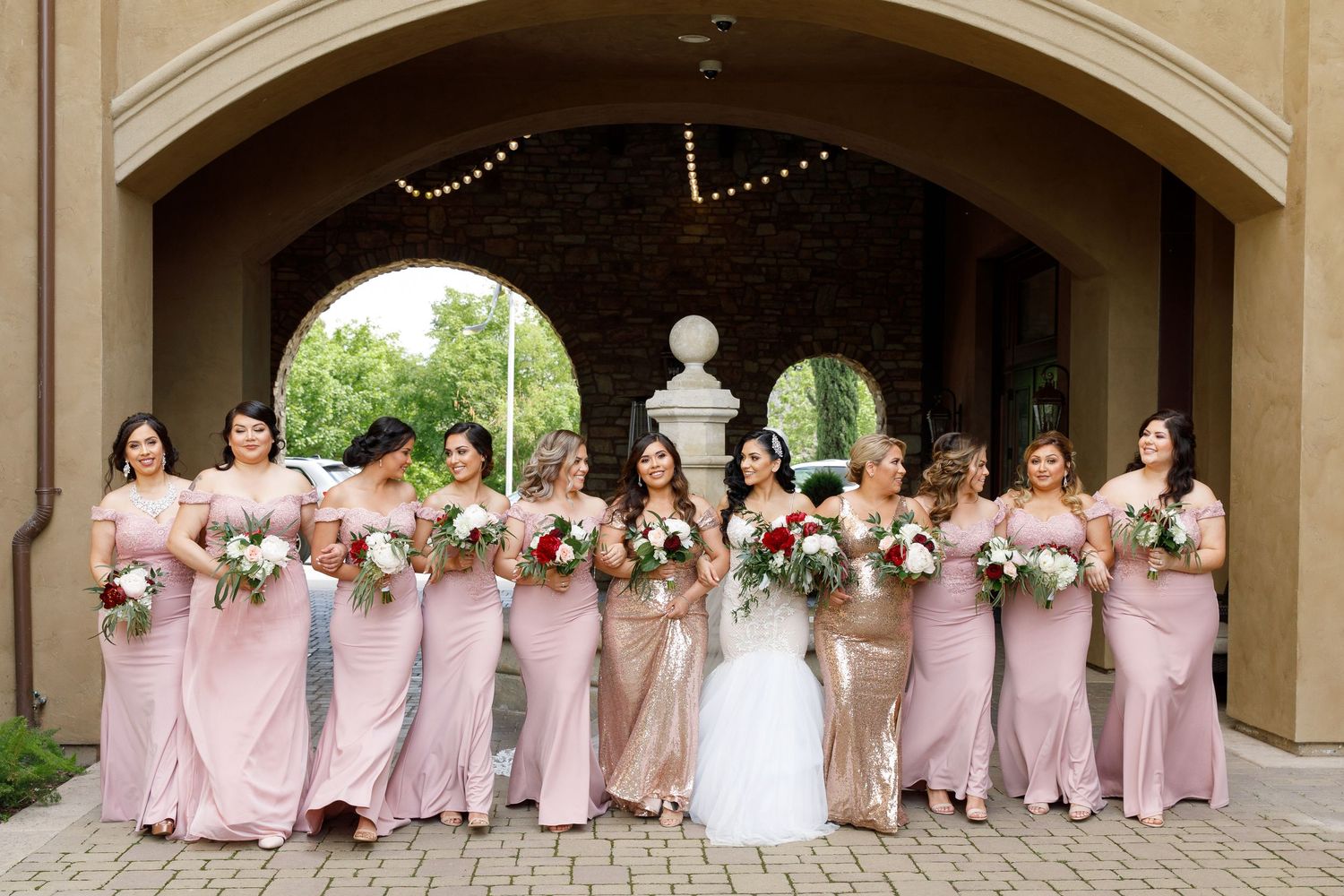 Wedding party in pink bridesmaid dresses poses under an elegant stone archway.