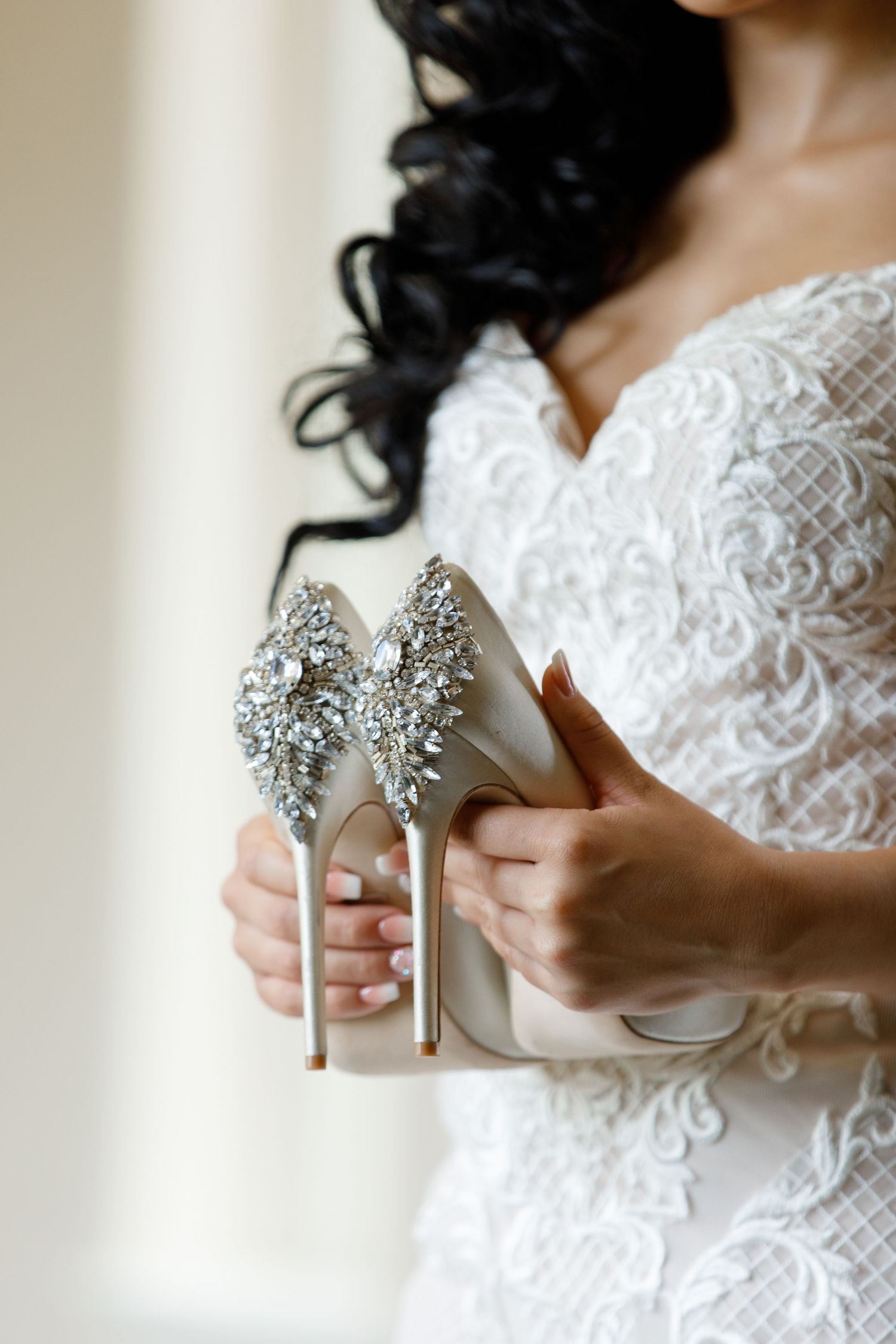 Elegant silver rhinestone bridal heels being held against a white lace dress background.