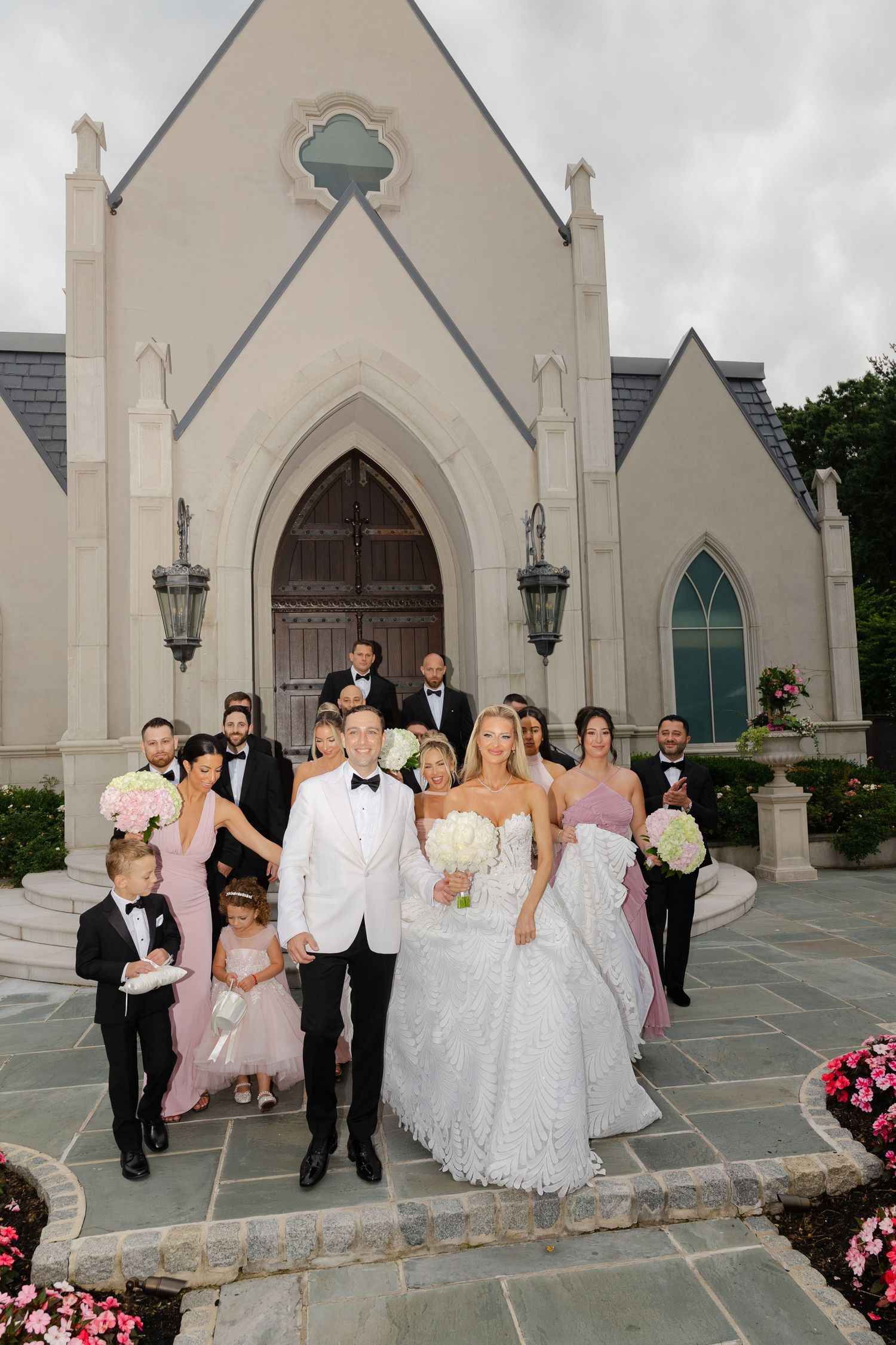 Wedding party poses for formal photos outside a beautiful gothic-style church with stone walls and arched doorways.