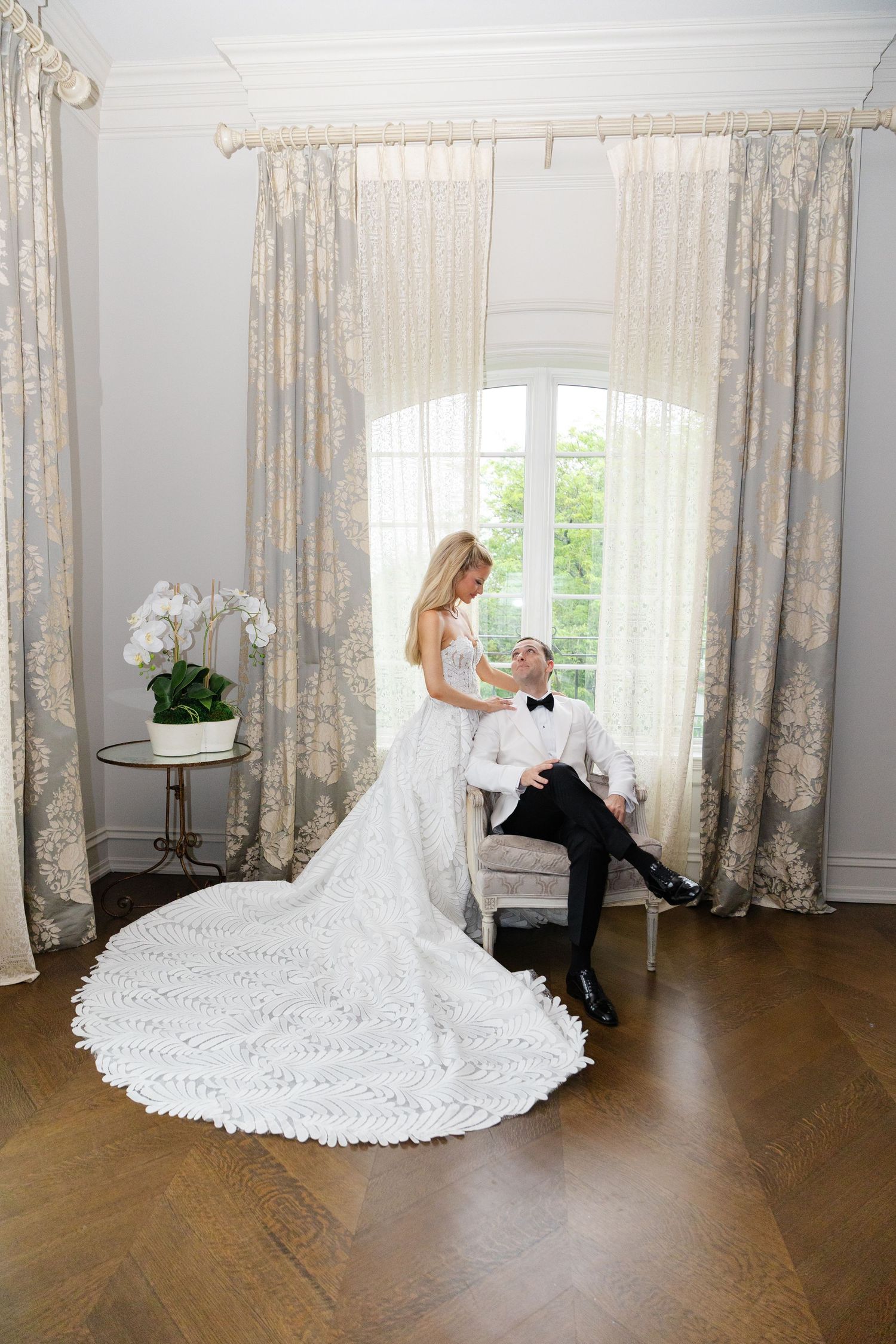 A bride in a flowing white wedding dress with a long train poses in an elegant room with floral curtains and hardwood floors.